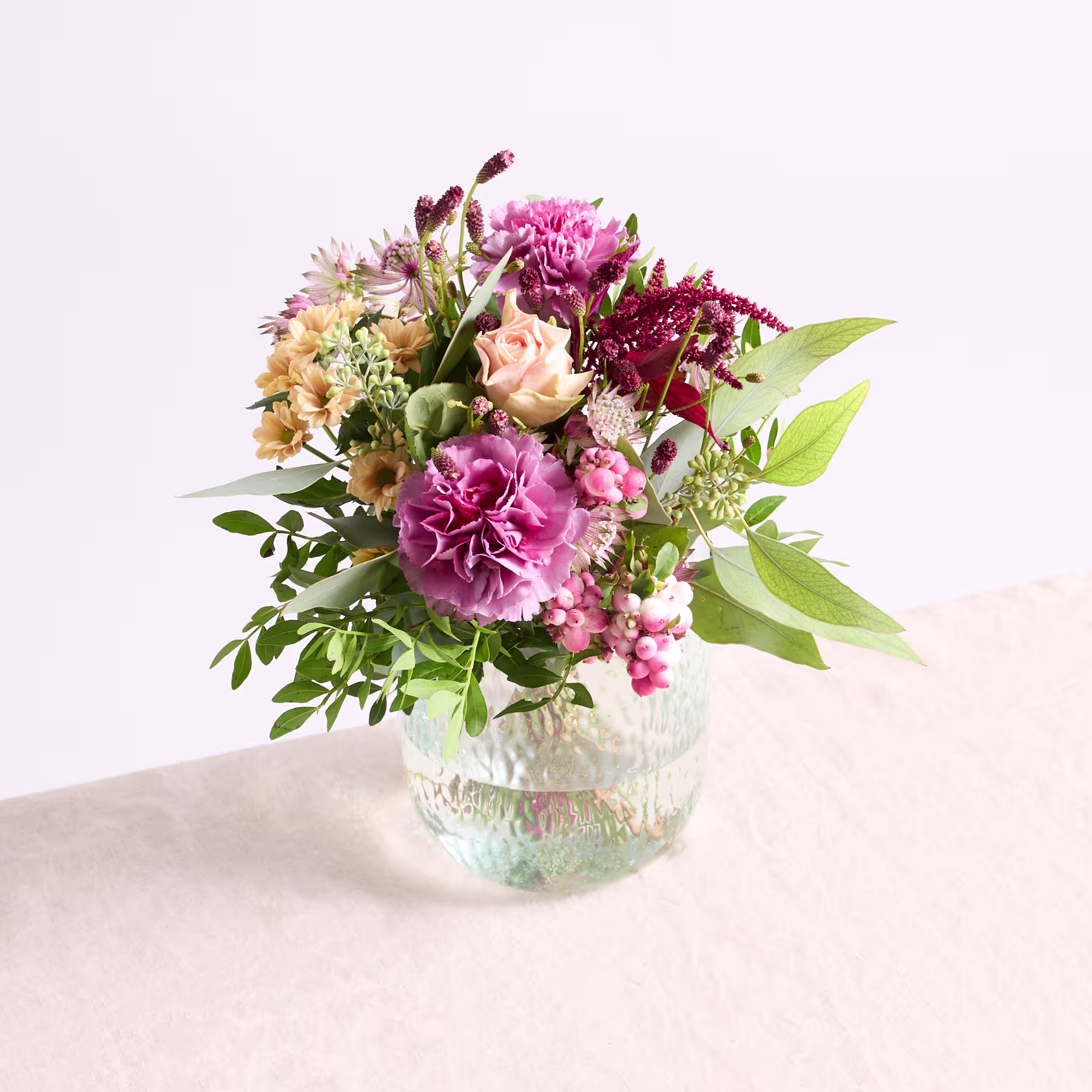 Ein leuchtender Strauß aus rosa Pfingstrosen, gelben Narzissen und anderen bunten Blumen in einer Glasvase steht auf einer hellen Fläche vor einem schlichten Hintergrund. (Beschreibungstext KI-generiert)