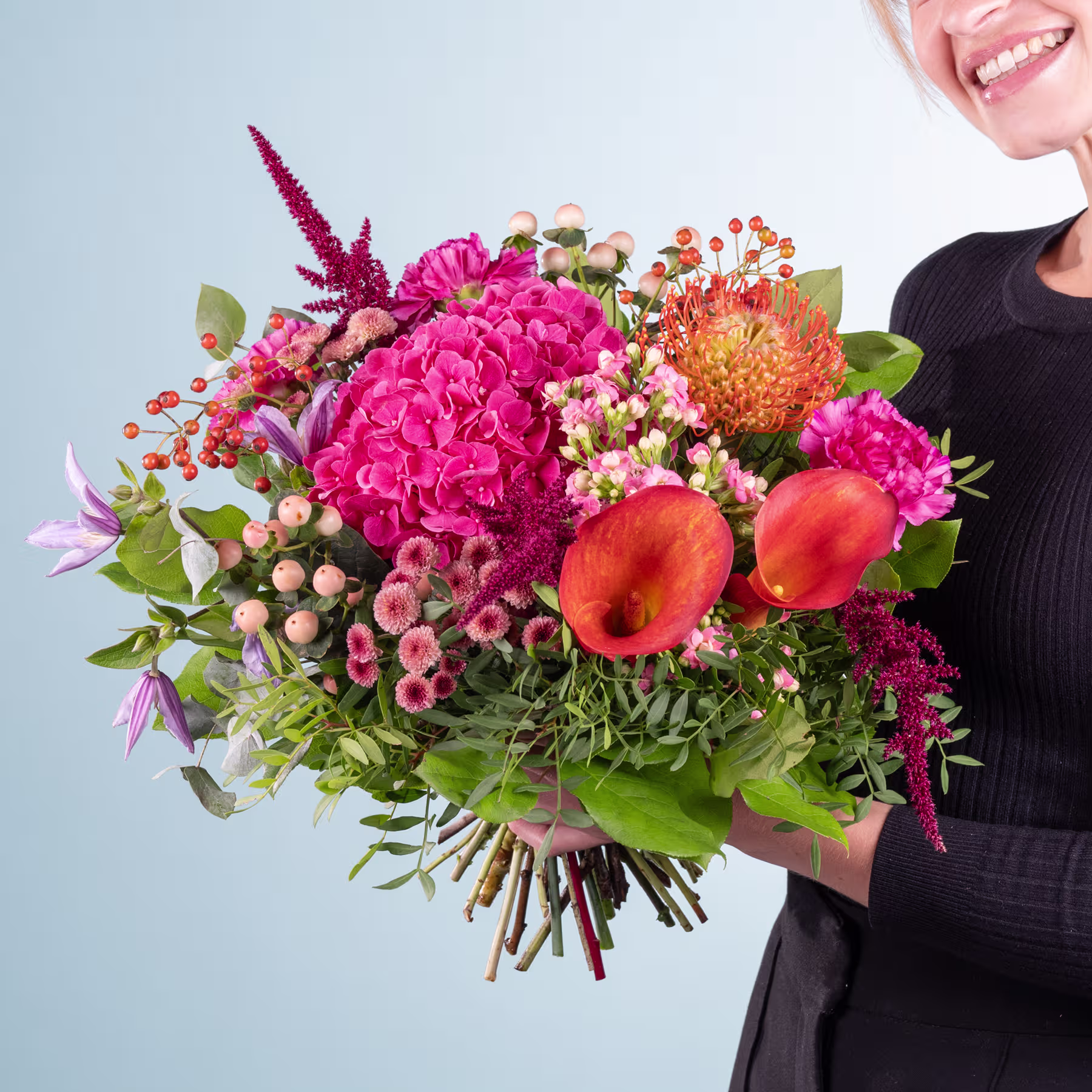 Ein leuchtender Strauß bunter Blumen, darunter rosa Hortensien, orangefarbene Proteen und verschiedene andere Blüten, gehalten von einer Person in einem schwarzen Outfit vor einem schlichten Hintergrund. (Beschreibungstext KI-generiert)