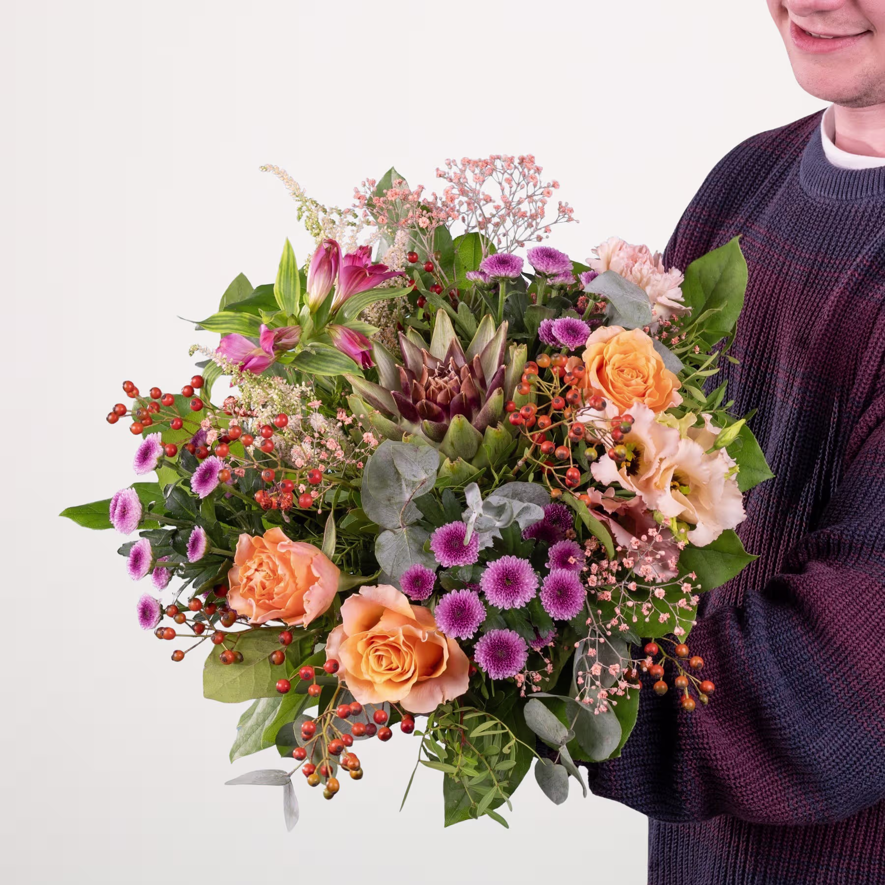 Ein lebendiger und farbenfroher Strauß verschiedener Blumen, darunter Rosen, Proteas und Gänseblümchen, wird von einer Person in einem dunklen Pullover vor einem schlichten weißen Hintergrund gehalten. (Beschreibungstext KI-generiert)