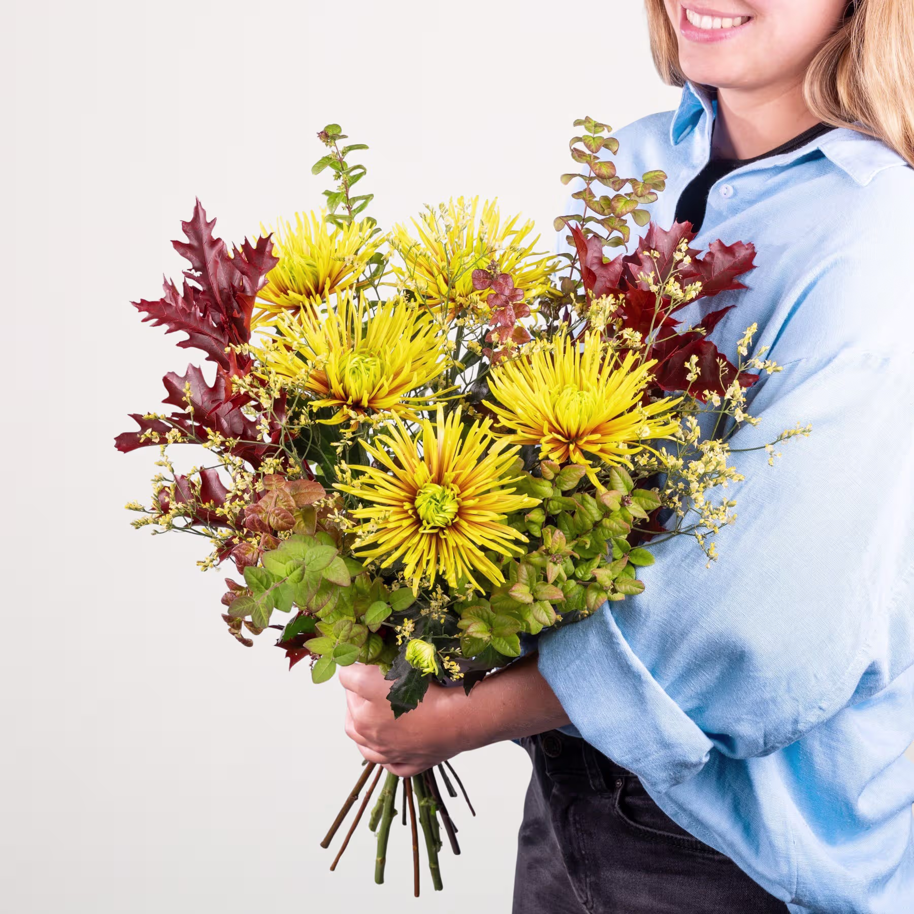 Ein Strauß leuchtend gelber Chrysanthemen und roter Herbstblätter wird von einer lächelnden Frau in einem blauen Hemd vor einem schlichten weißen Hintergrund gehalten. (Beschreibungstext KI-generiert)