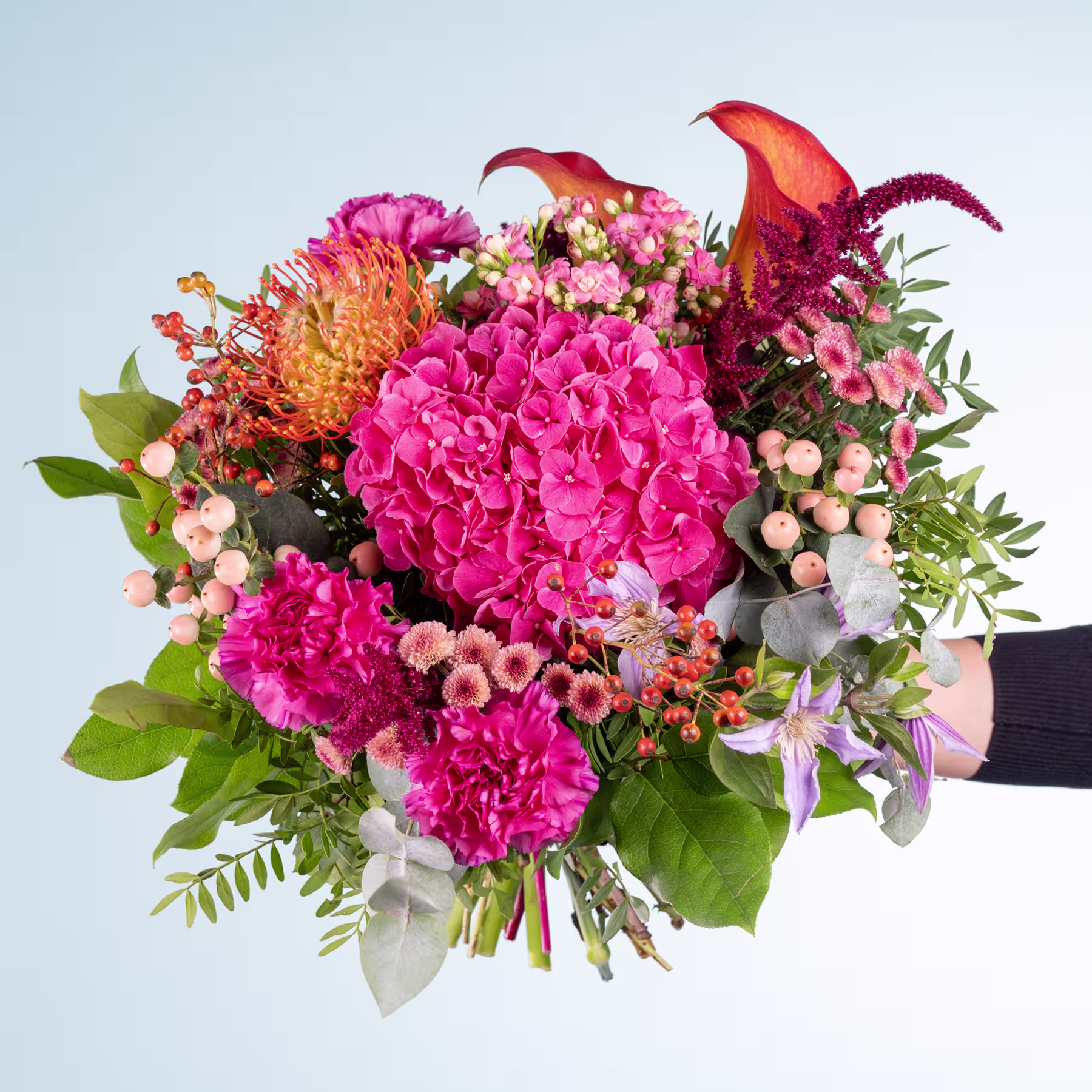 Ein lebendiger Strauß aus rosa Hortensien, orangefarbenen Dahlien und anderen bunten Blumen vor einem schlichten blauen Hintergrund. (Beschreibungstext KI-generiert)
