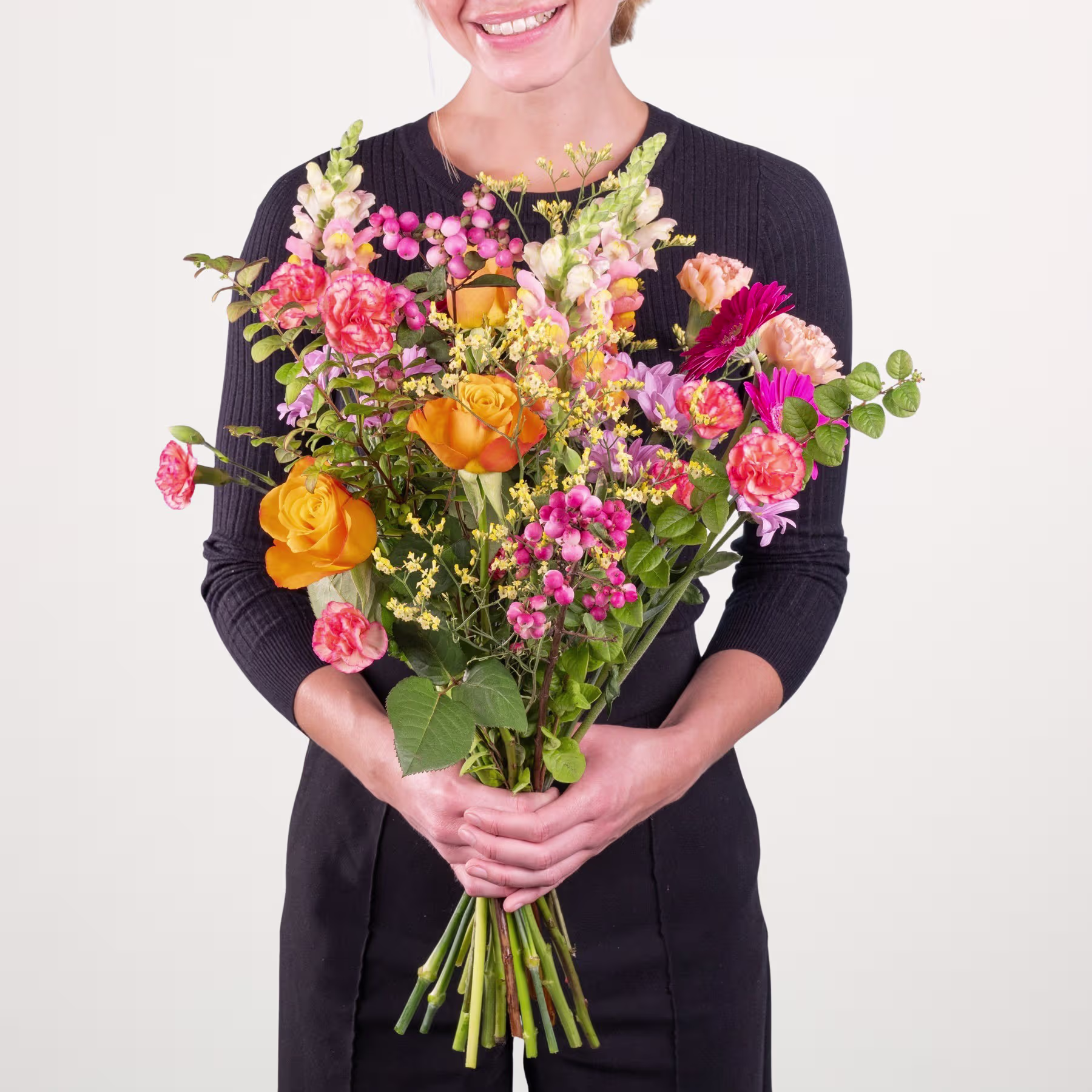 Eine lächelnde Frau in einem schwarzen Oberteil hält vor einem schlichten weißen Hintergrund einen leuchtenden Strauß bunter Blumen, darunter Rosen, Nelken und andere Blüten. (Beschreibungstext KI-generiert)