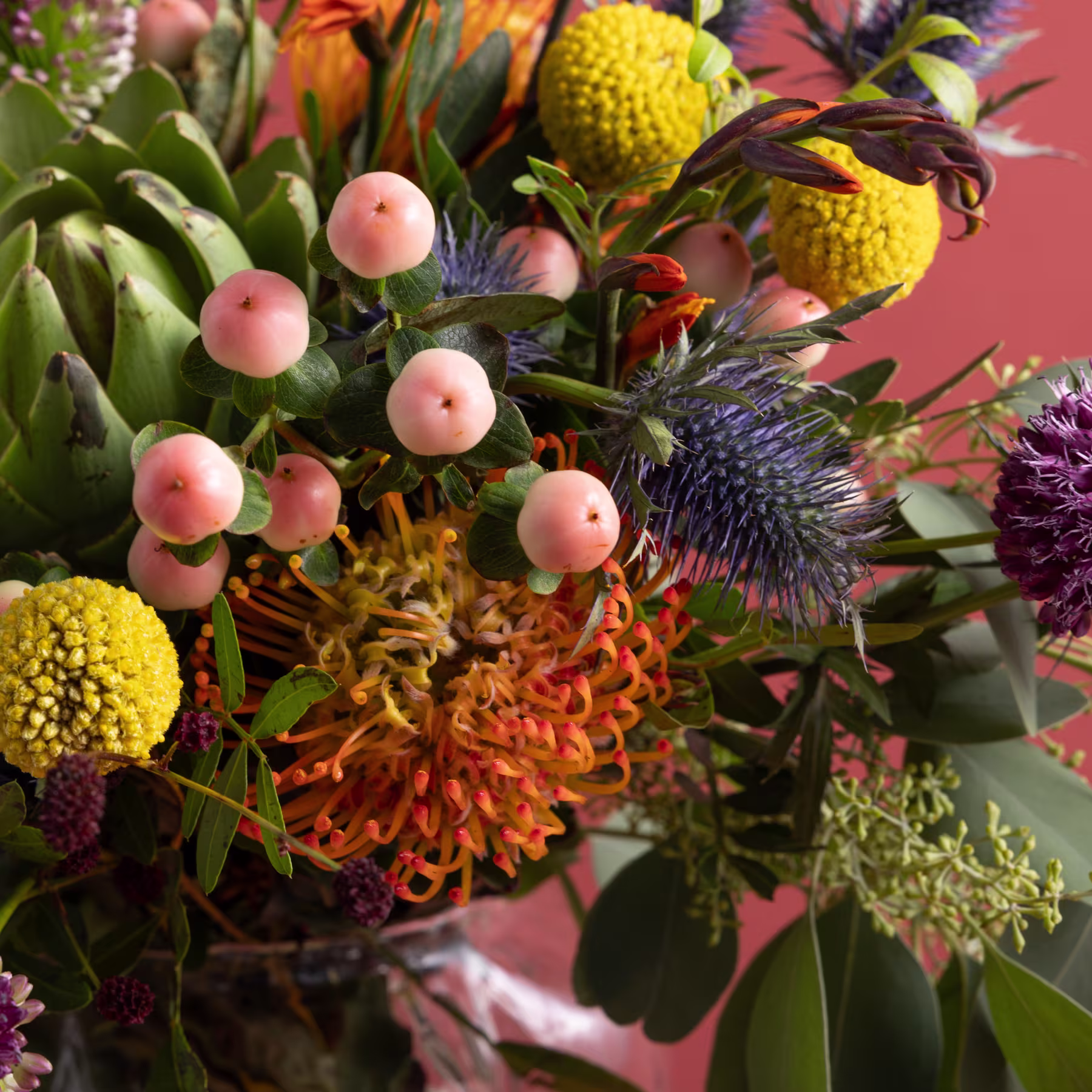 Ein lebendiger Strauß bunter Blumen, darunter gelbe Chrysanthemen, rosa Beeren und eine auffällige violette Distel, vor einem zartrosa Hintergrund. (Beschreibungstext KI-generiert)