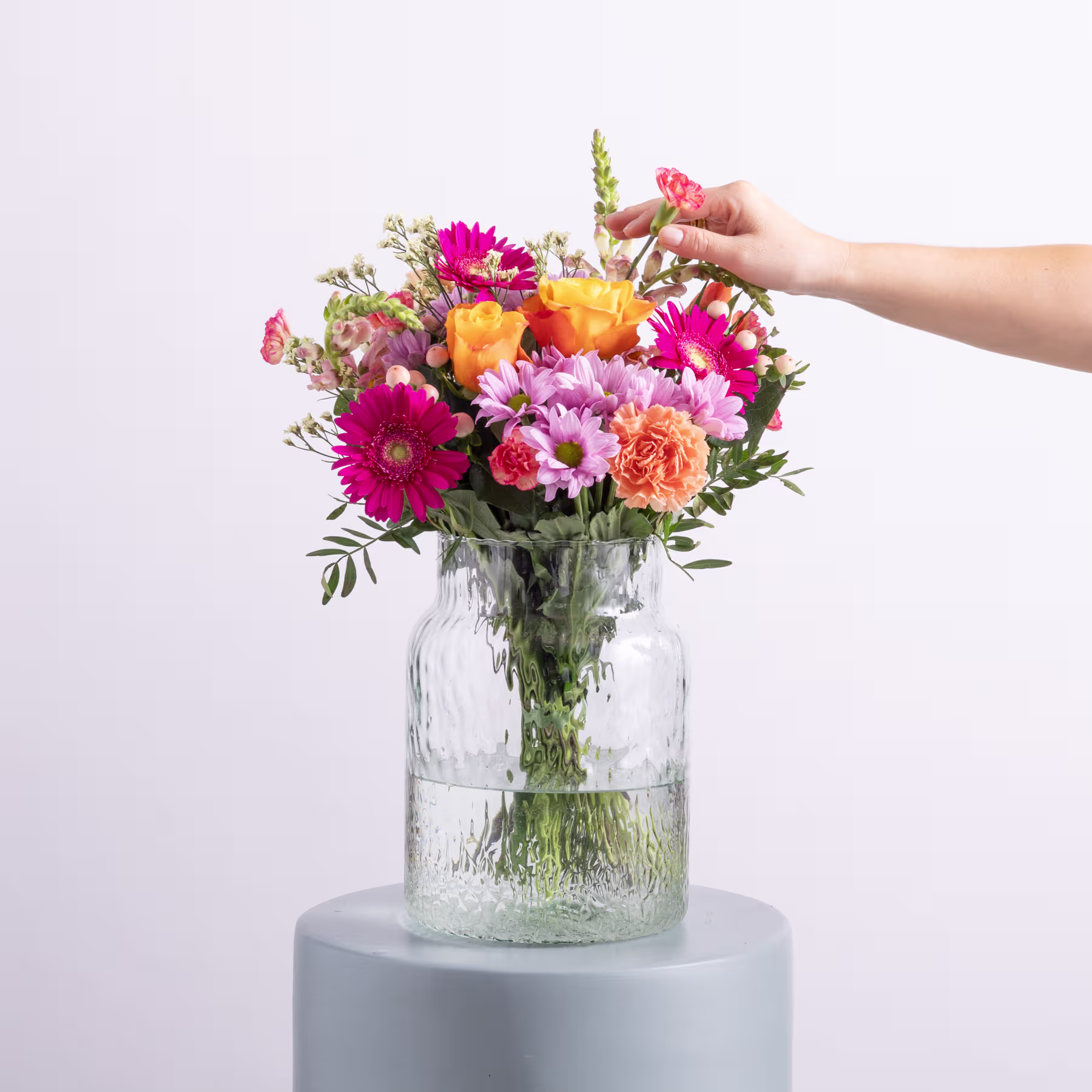 Ein lebendiger Strauß bunter Blumen, darunter Gerbera, Nelken und andere Blüten, ist in einer Glasvase vor einem schlichten weißen Hintergrund arrangiert. (Beschreibungstext KI-generiert)