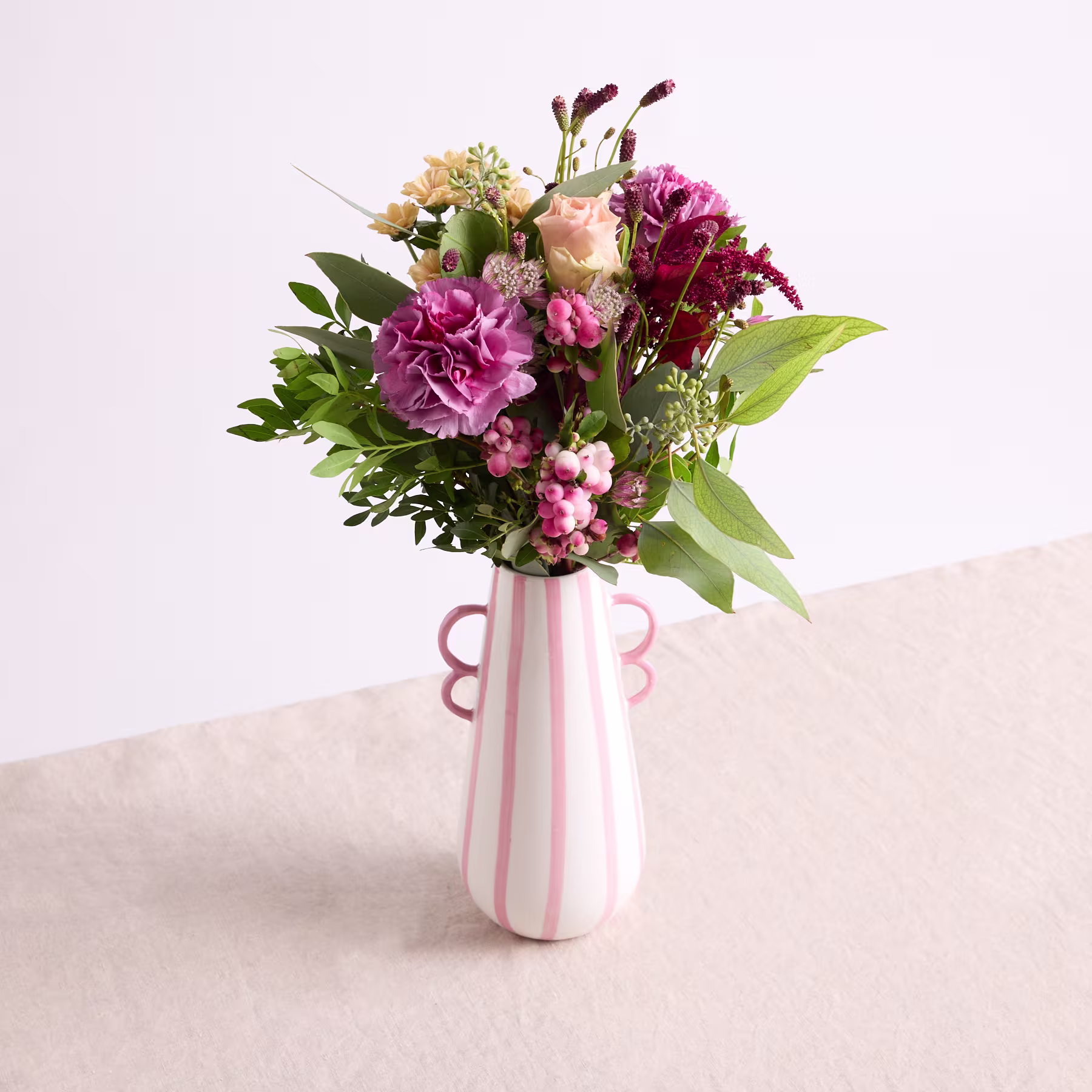 Ein farbenfroher Strauß rosa und weißer Blumen, darunter Pfingstrosen und Nelken, wird in einer rosa und weiß gestreiften Vase vor einem schlichten Hintergrund präsentiert. (Beschreibungstext KI-generiert)
