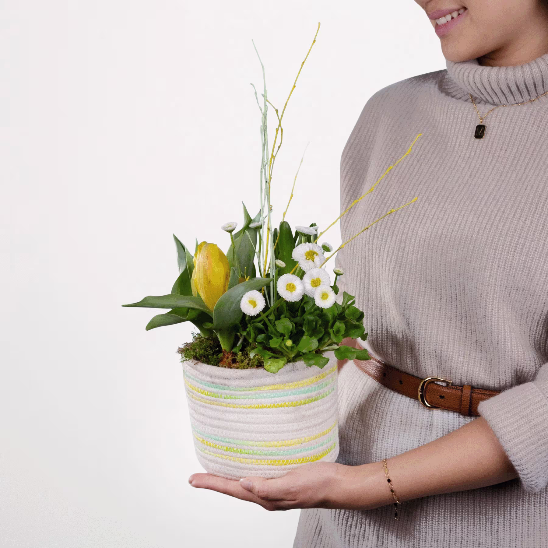 Eine Person hält eine Topfpflanze mit gelben Tulpen und weißen Gänseblümchen vor einem schlichten weißen Hintergrund. (Beschreibungstext KI-generiert)
