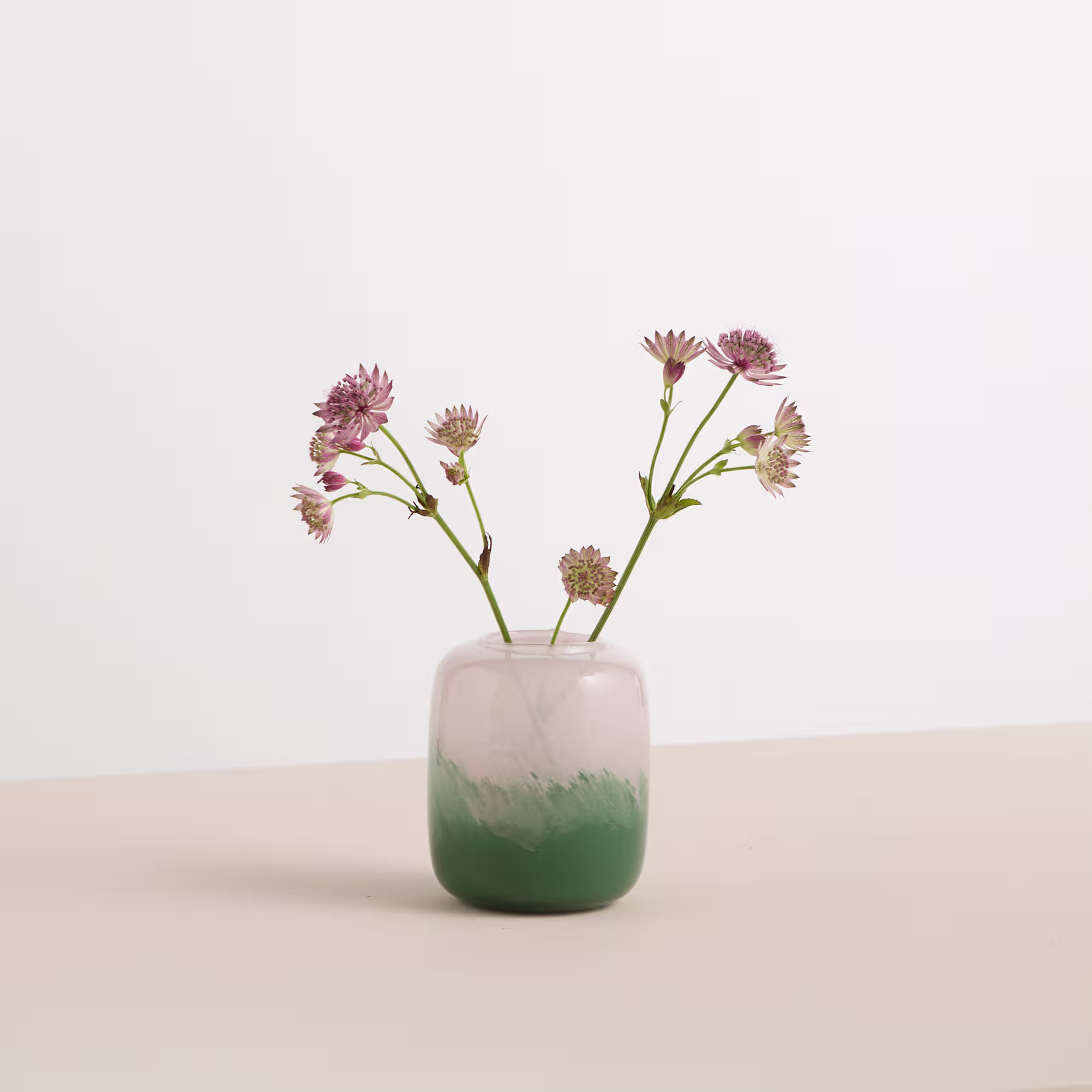 Eine Vase mit rosa und violetten Blumen steht auf einer Holzoberfläche vor einem schlichten weißen Hintergrund. (Beschreibungstext KI-generiert)