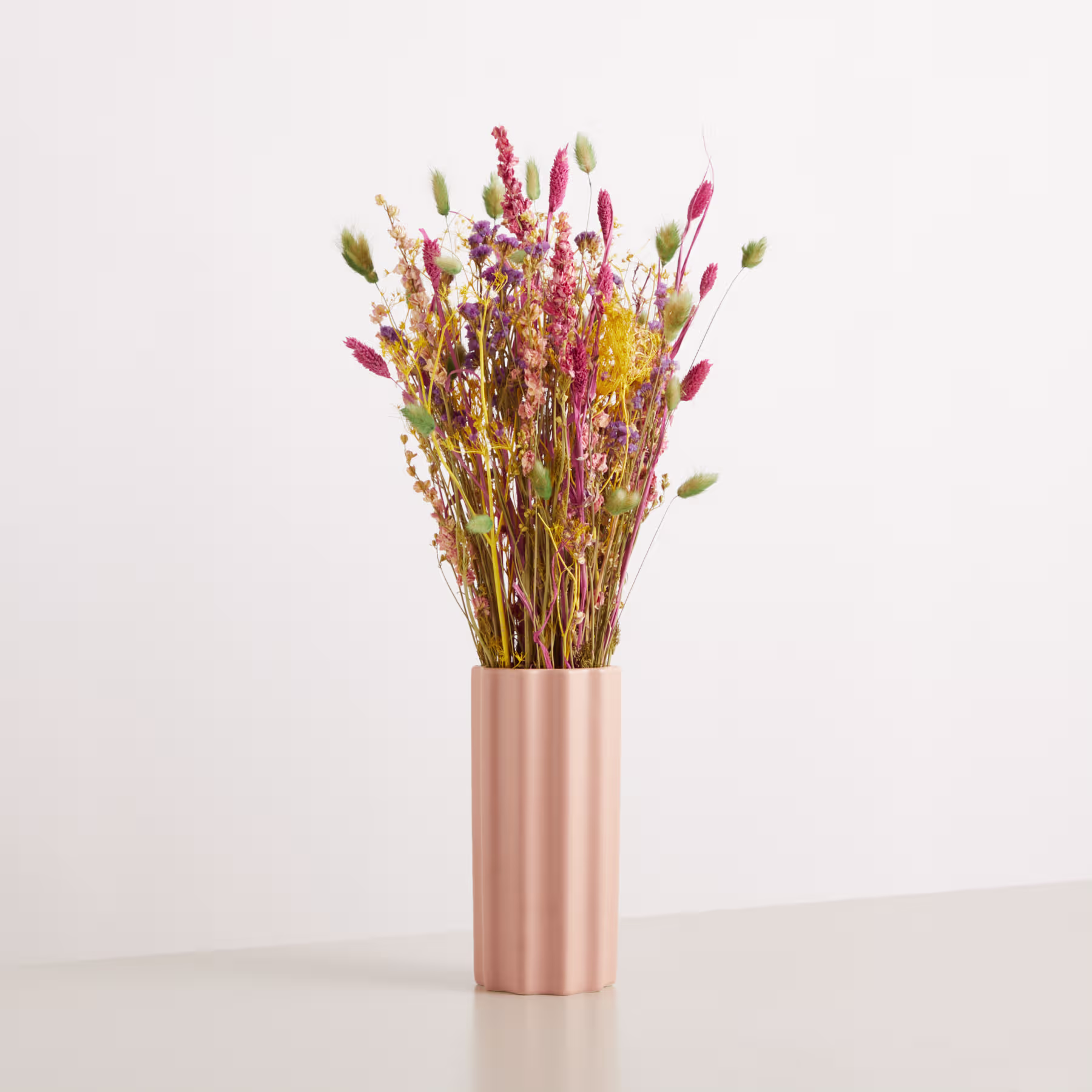 Ein Strauß leuchtender Trockenblumen in einer metallisch rosa Vase steht vor einem schlichten weißen Hintergrund. (Beschreibungstext KI-generiert)