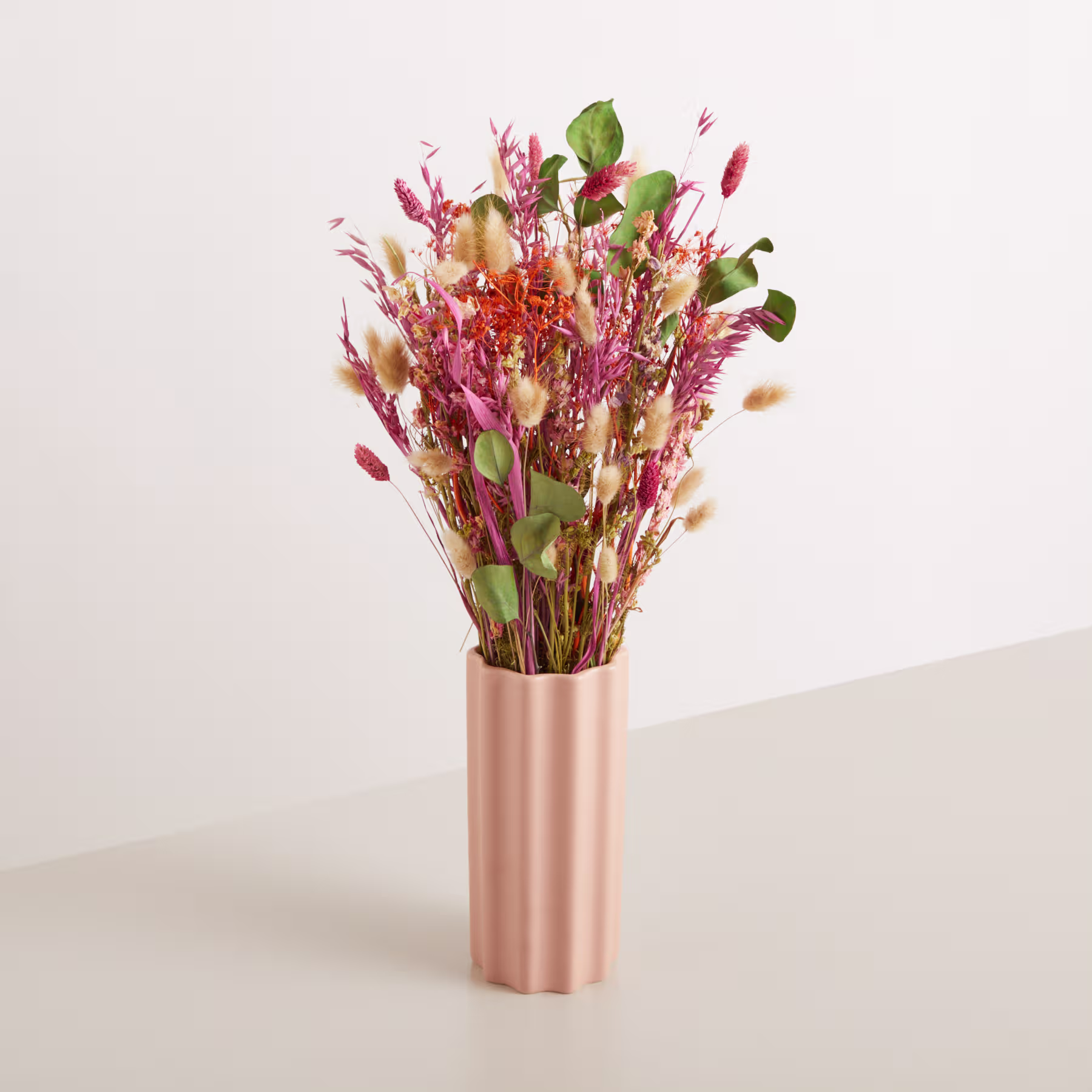 Ein farbenfroher Strauß getrockneter Blumen in einer metallisch rosa Vase steht vor einem schlichten, hellen Hintergrund. (Beschreibungstext KI-generiert)