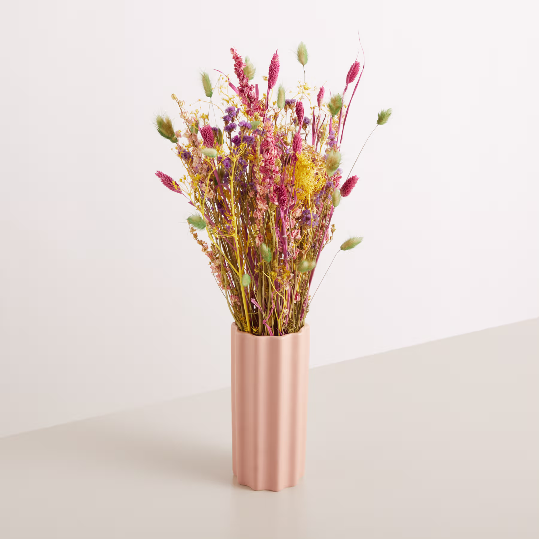 Ein Strauß leuchtender Trockenblumen in verschiedenen Rosa-, Rot- und Gelbtönen wird in einer hohen, zylindrischen Vase in blassem Rosa vor einem schlichten, hellen Hintergrund präsentiert. (Beschreibungstext KI-generiert)