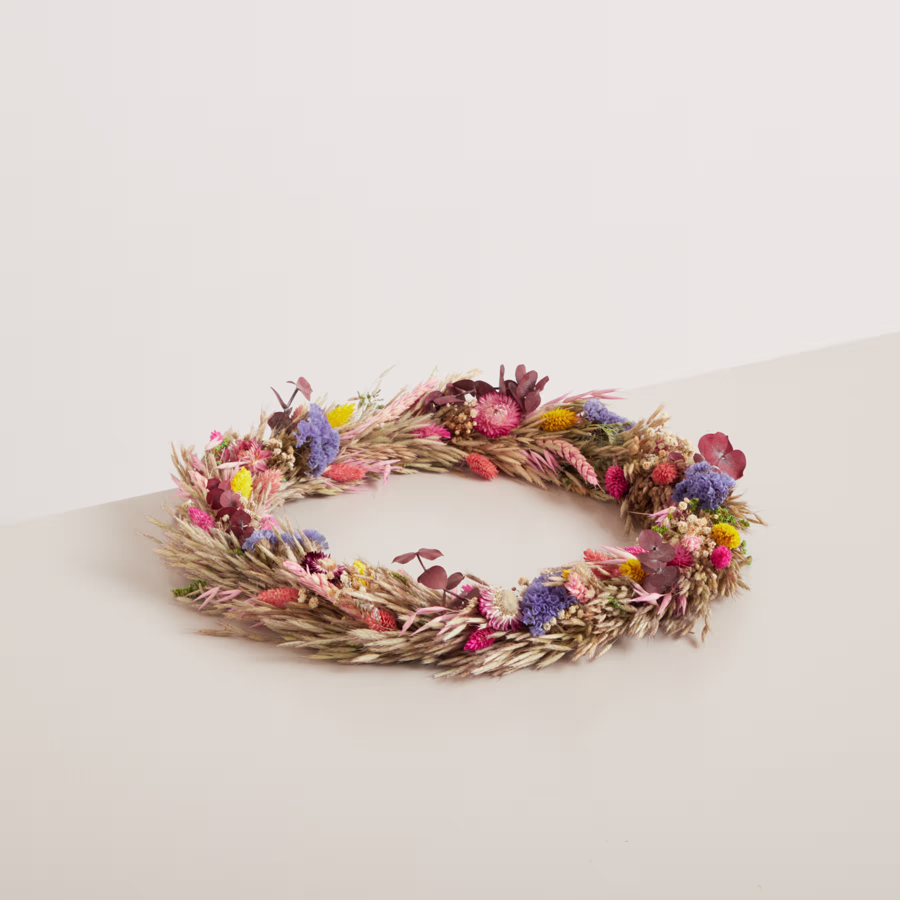 Auf einer schlichten, hellen Oberfläche liegt ein bunter Kranz aus getrockneten Blumen, Blättern und anderen Naturelementen. (Beschreibungstext KI-generiert)