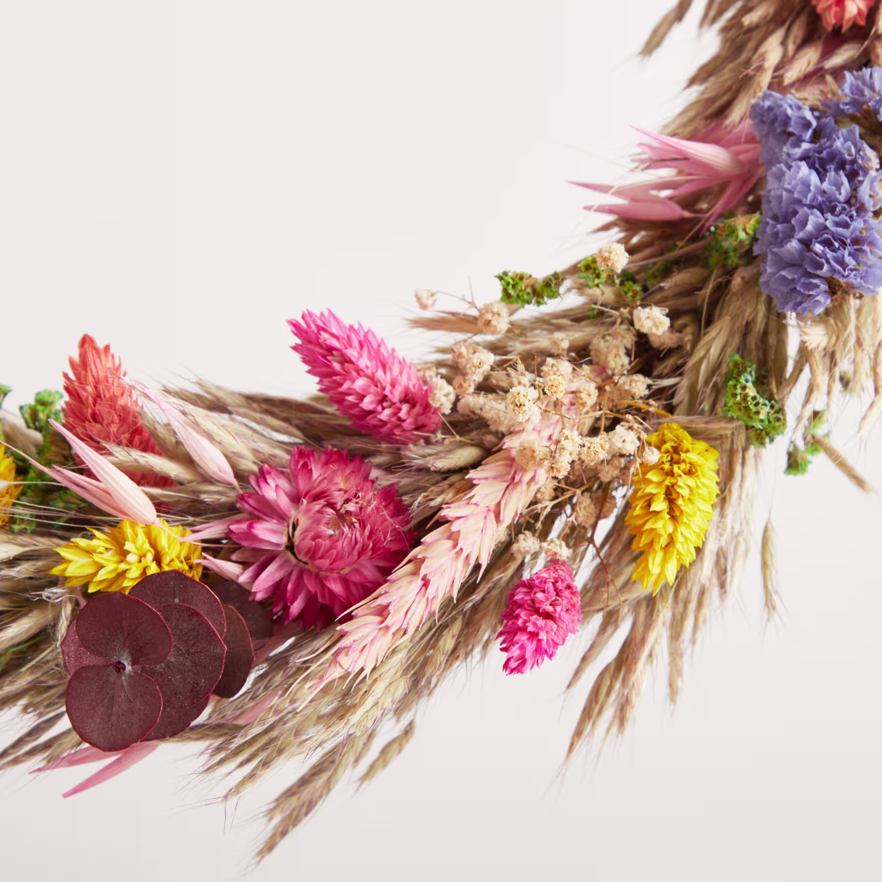 Ein lebendiger Strauß getrockneter Blumen, darunter rosa und violette Chrysanthemen, Weizenhalme und andere natürliche Elemente, vor einem schlichten weißen Hintergrund. (Beschreibungstext KI-generiert)