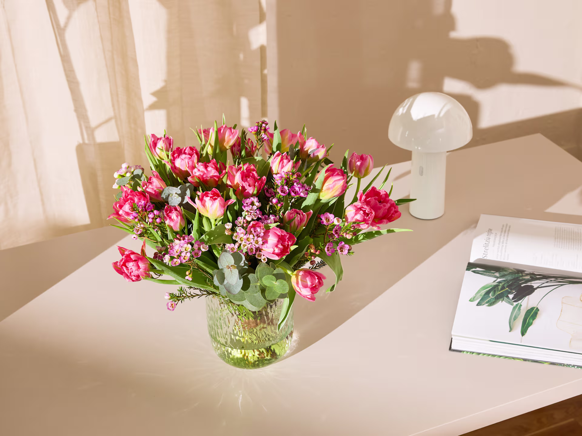 Ein farbenfroher Strauß rosa Tulpen und Nelken steht auf einem Holztisch, im Hintergrund eine Lampe und ein gerahmtes Foto, was eine gemütliche und einladende Atmosphäre schafft. (Beschreibungstext KI-generiert)