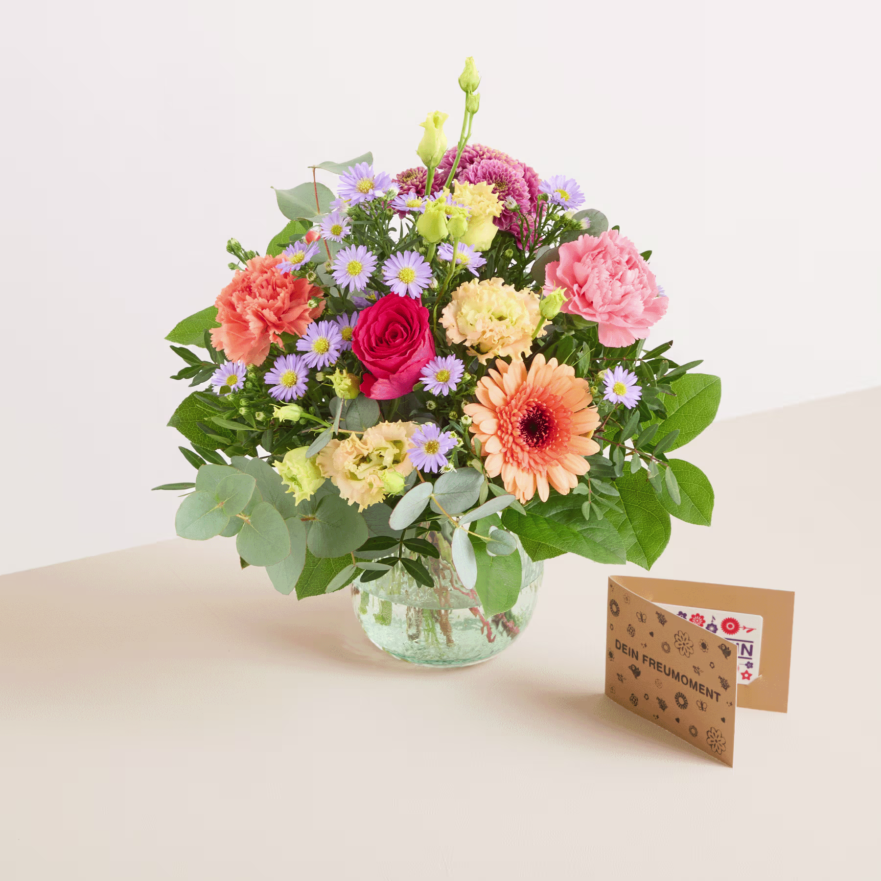 Ein lebendiger Strauß bunter Blumen, darunter Rosen, Gerbera und Nelken, arrangiert in einer Glasvase auf einer Holzoberfläche, mit einer Grußkarte davor. (Beschreibungstext KI-generiert)