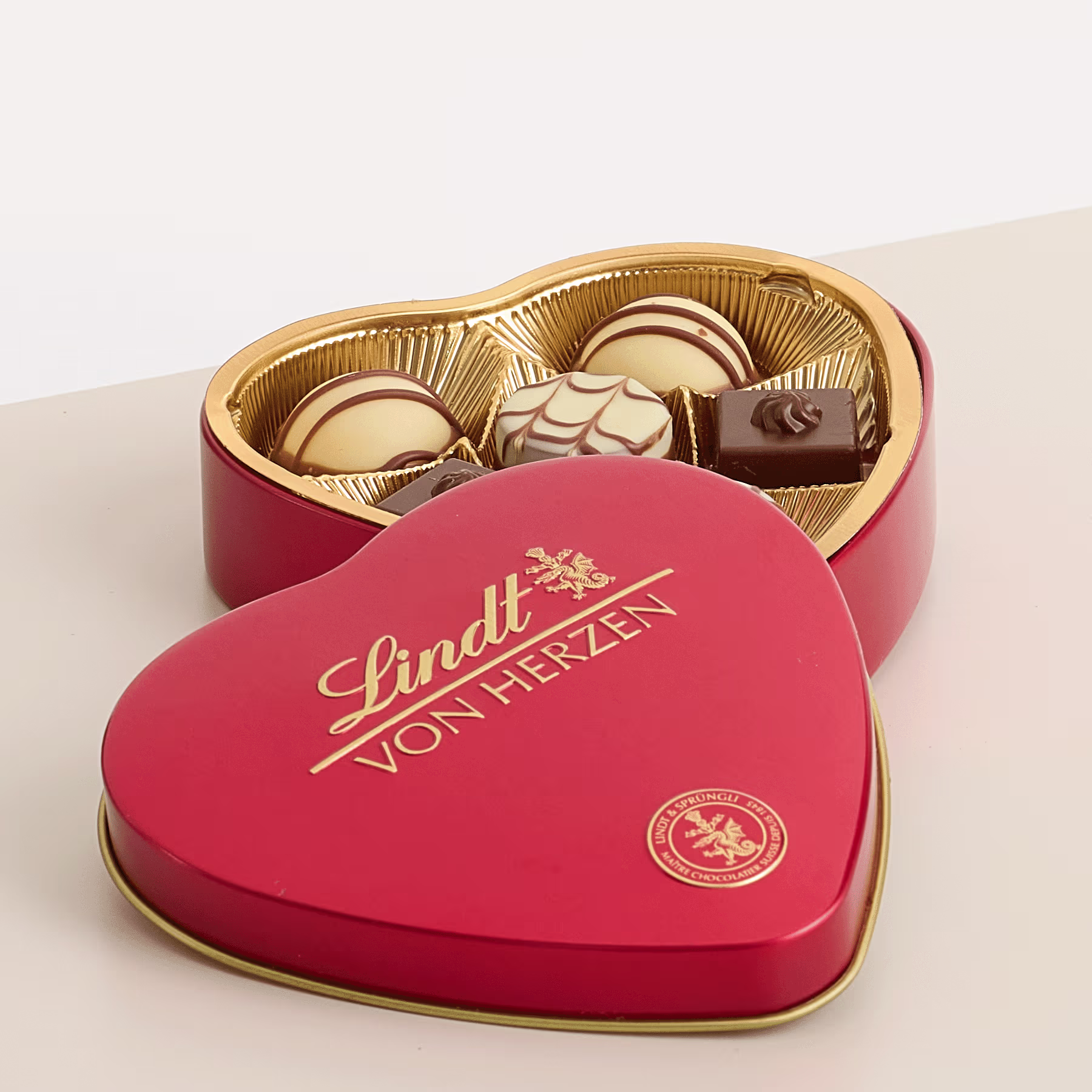 Eine herzförmige rote Schachtel mit einer Auswahl an Lindt-Schokolade vor einem schlichten Hintergrund. (Beschreibungstext KI-generiert)