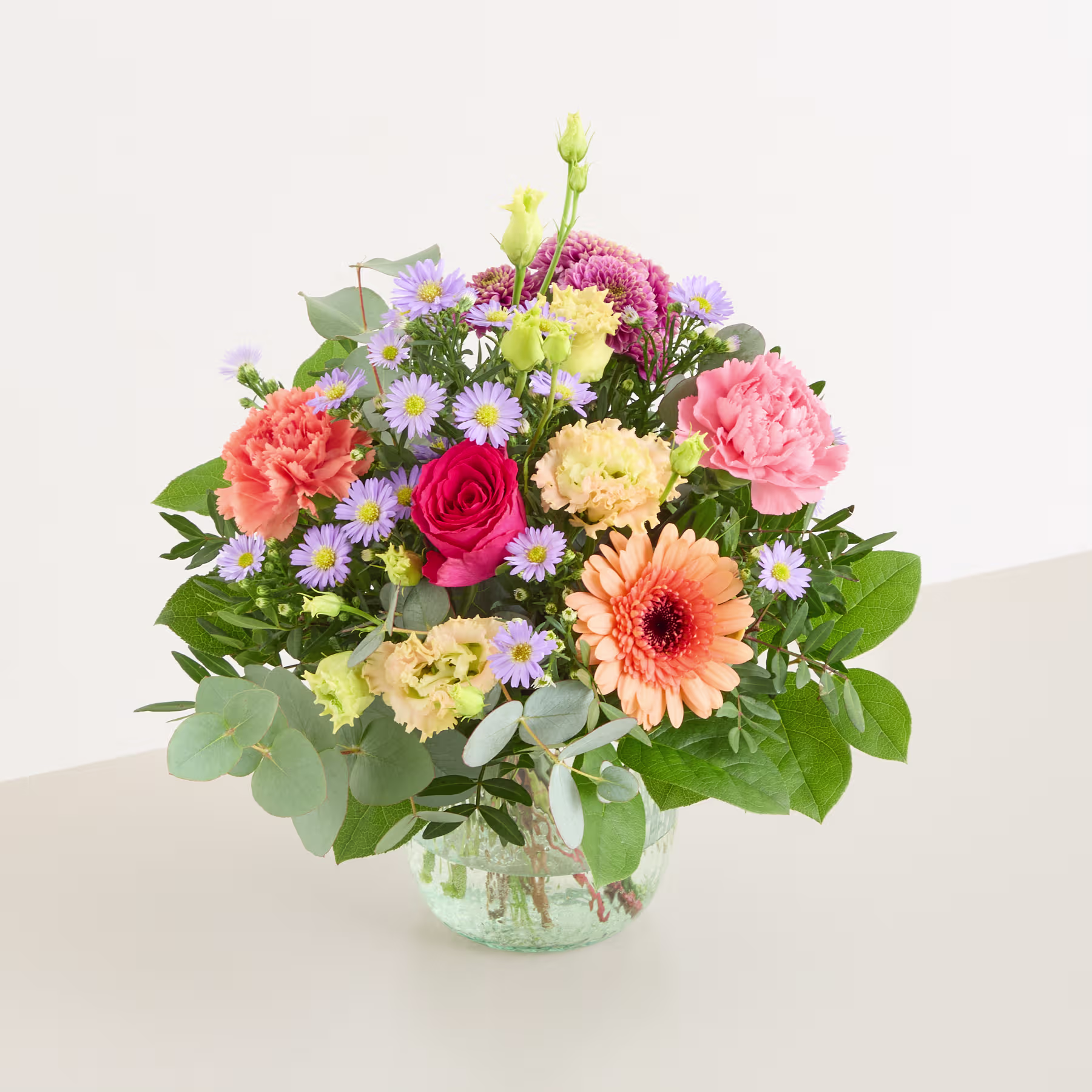 Ein lebendiger Strauß bunter Blumen, darunter Rosen, Gerbera und Chrysanthemen, arrangiert in einer Glasvase vor einem schlichten weißen Hintergrund. (Beschreibungstext KI-generiert)