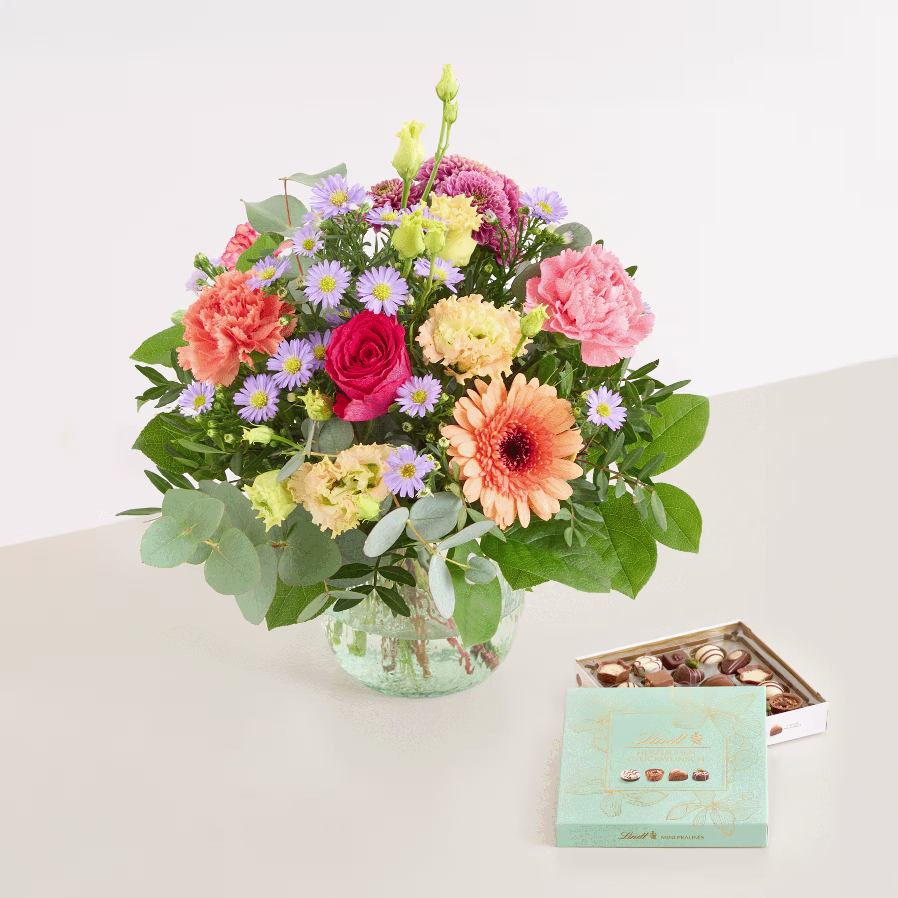 Ein farbenfroher Strauß bunter Blumen, darunter Rosen, Gerbera und Chrysanthemen, steht auf einem Tisch neben einer Schachtel Pralinen. (Beschreibungstext KI-generiert)