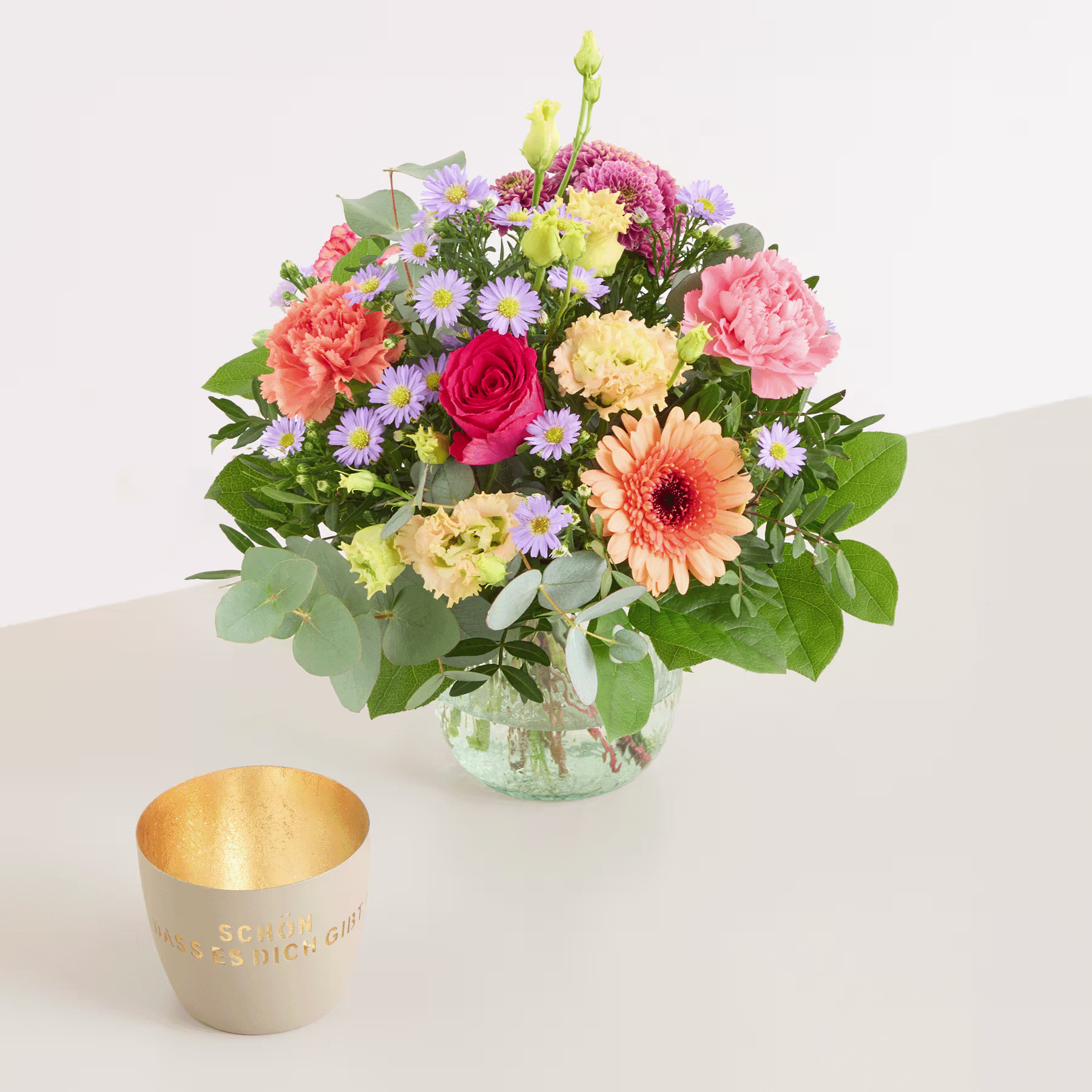 Ein leuchtender Strauß bunter Blumen, darunter Rosen, Nelken und Gänseblümchen, wird in einer Glasvase auf einem Tisch präsentiert, begleitet von einem goldenen Kerzenhalter. (Beschreibungstext KI-generiert)