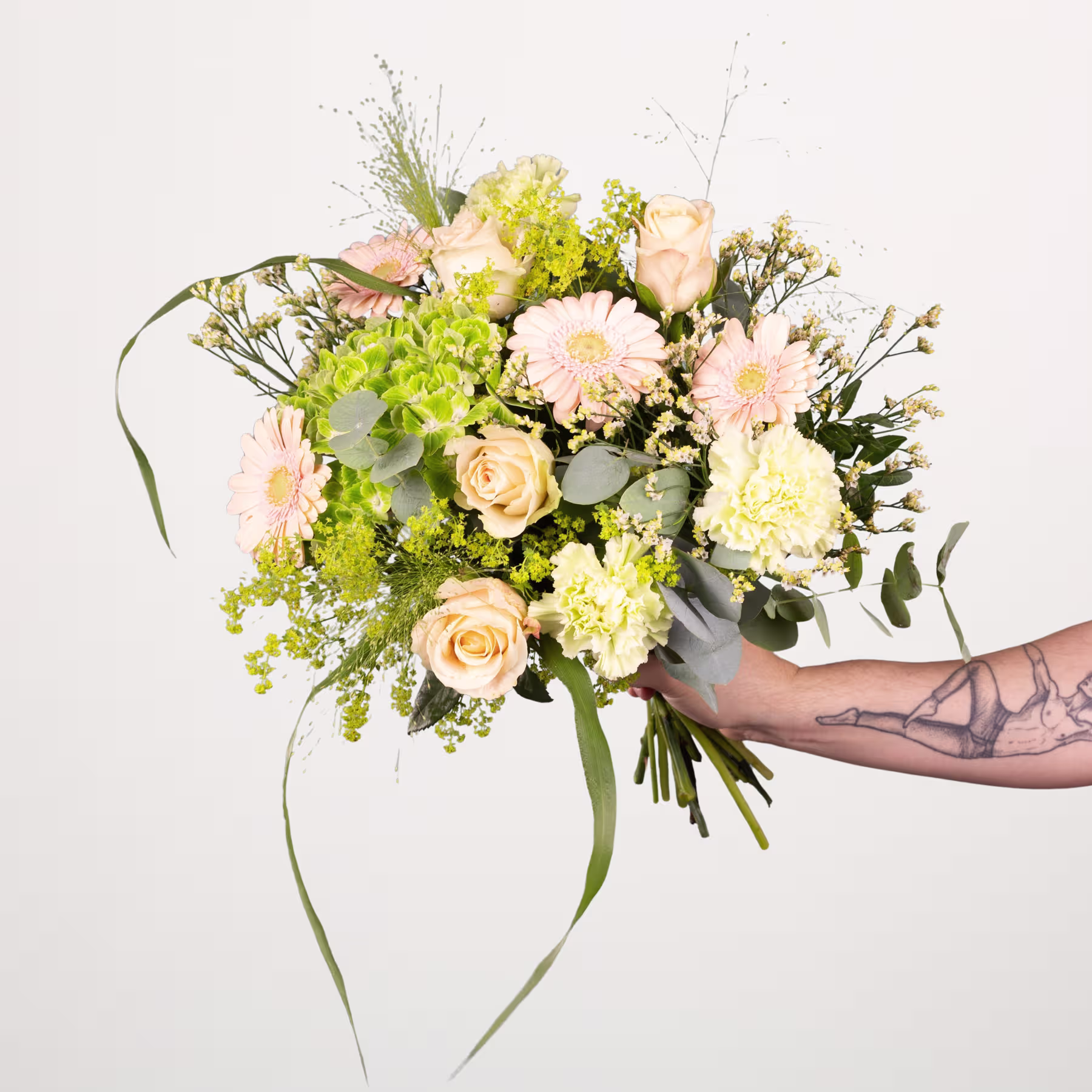 Ein farbenfroher Strauß verschiedener Blumen, darunter Rosen, Gänseblümchen und Grünpflanzen, wird in einer tätowierten Hand vor einem schlichten weißen Hintergrund gehalten. (Beschreibungstext KI-generiert)
