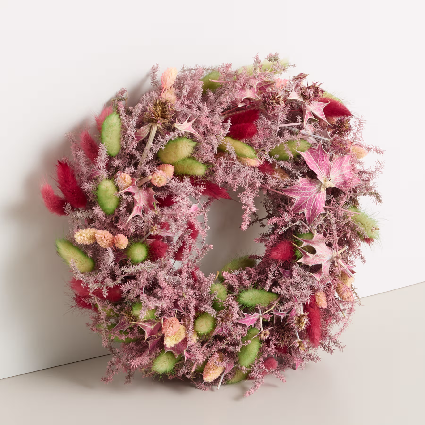 Ein leuchtender, runder Kranz aus Blättern in verschiedenen Rosa- und Grüntönen, geschmückt mit roten Beeren und anderen natürlichen Elementen, liegt auf einem schlichten weißen Hintergrund. (Beschreibungstext KI-generiert)