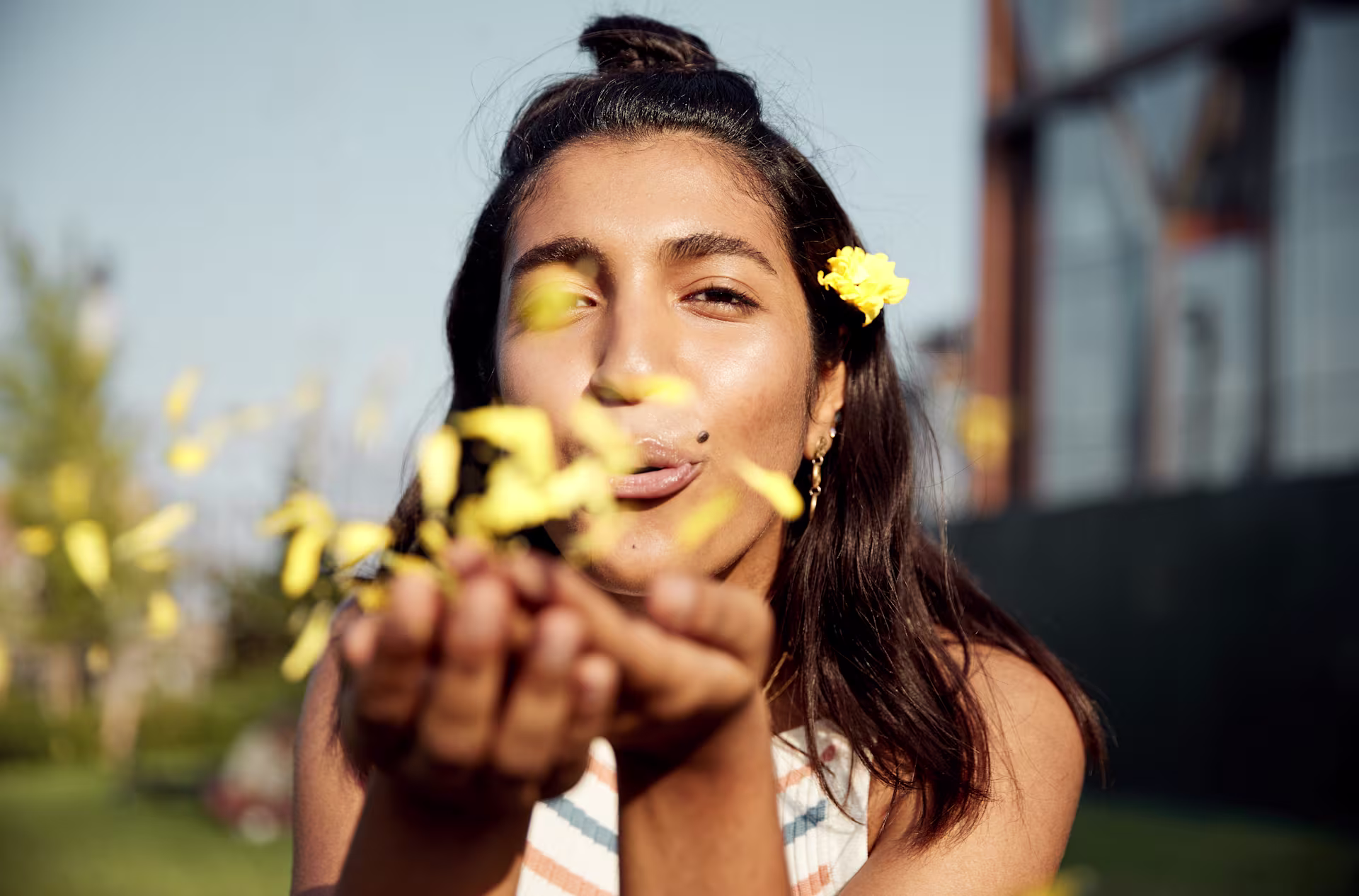 Eine junge Frau mit langem dunklem Haar und einer gelben Blume im Haar lächelt und hält eine Handvoll gelber Blumen vor einem unscharfen natürlichen Hintergrund hoch. (Beschreibungstext KI-generiert)