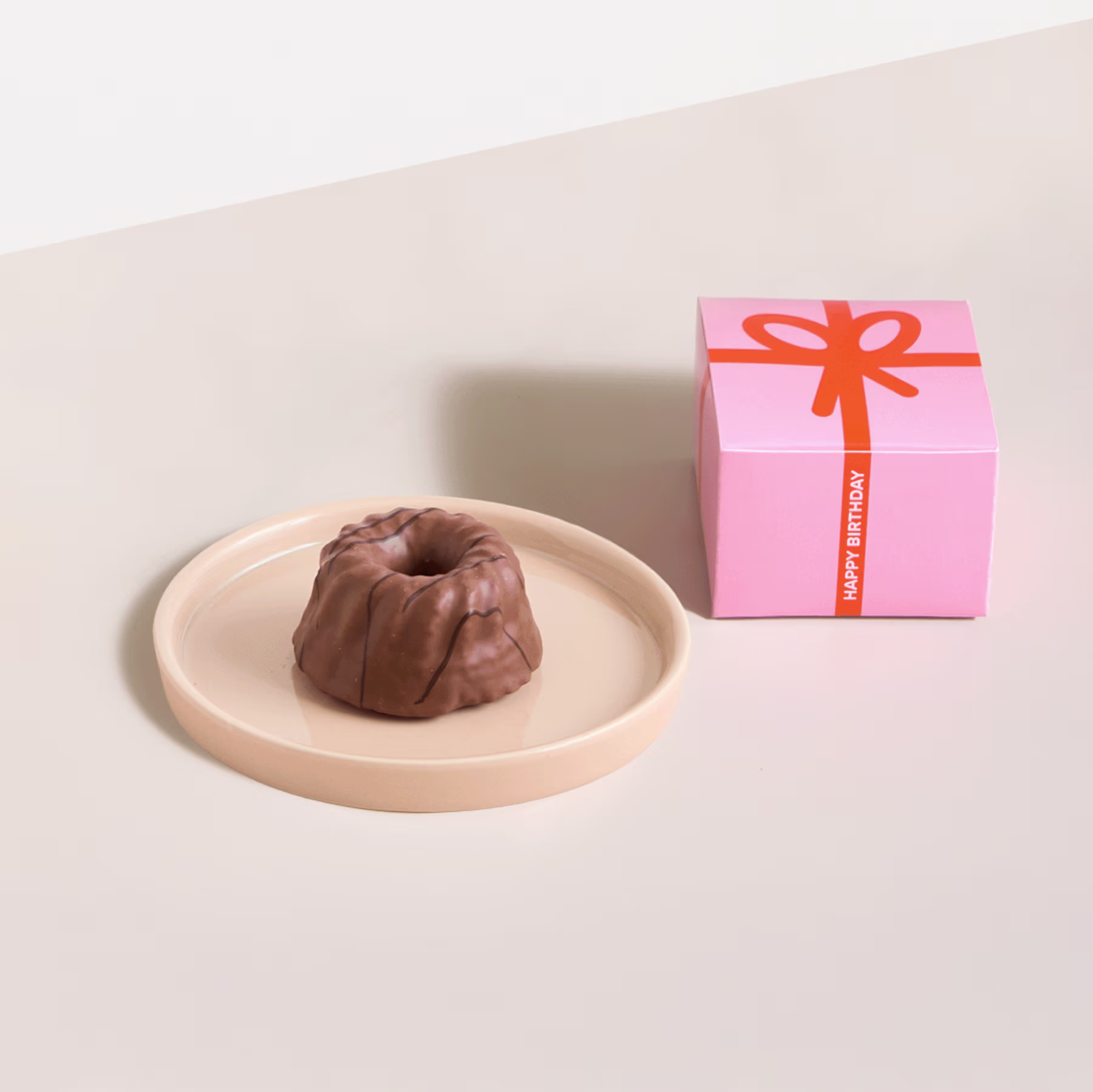 Eine rosa Geschenkbox mit orangefarbener Schleife steht neben einem runden Schokoladendessert auf einem hellen Teller. (Beschreibungstext KI-generiert)