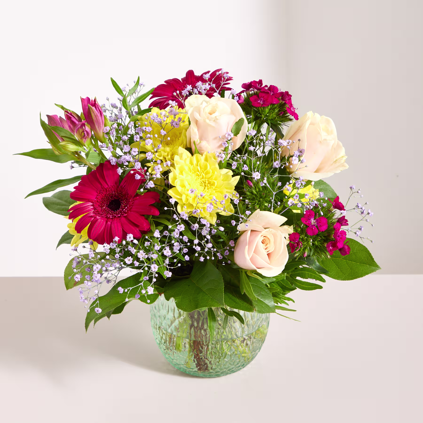 Ein lebendiger Strauß verschiedener bunter Blumen, darunter Rosen, Gerbera und andere Blüten, arrangiert in einer Glasvase vor einem schlichten Hintergrund. (Beschreibungstext KI-generiert)