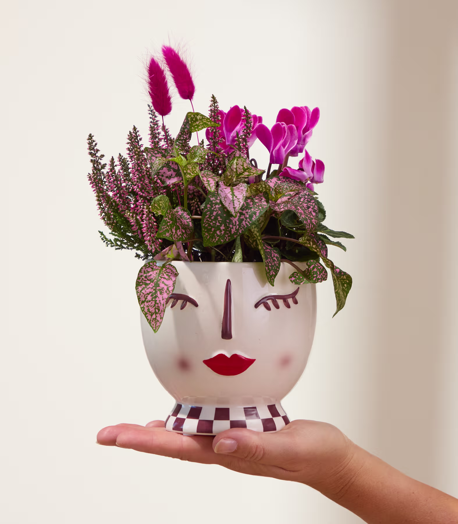 Ein skurriler Keramik-Übertopf in Form eines Gesichts mit geschlossenen Augen, geschmückt mit leuchtend rosa Blumen und Grünpflanzen. (Beschreibungstext KI-generiert)