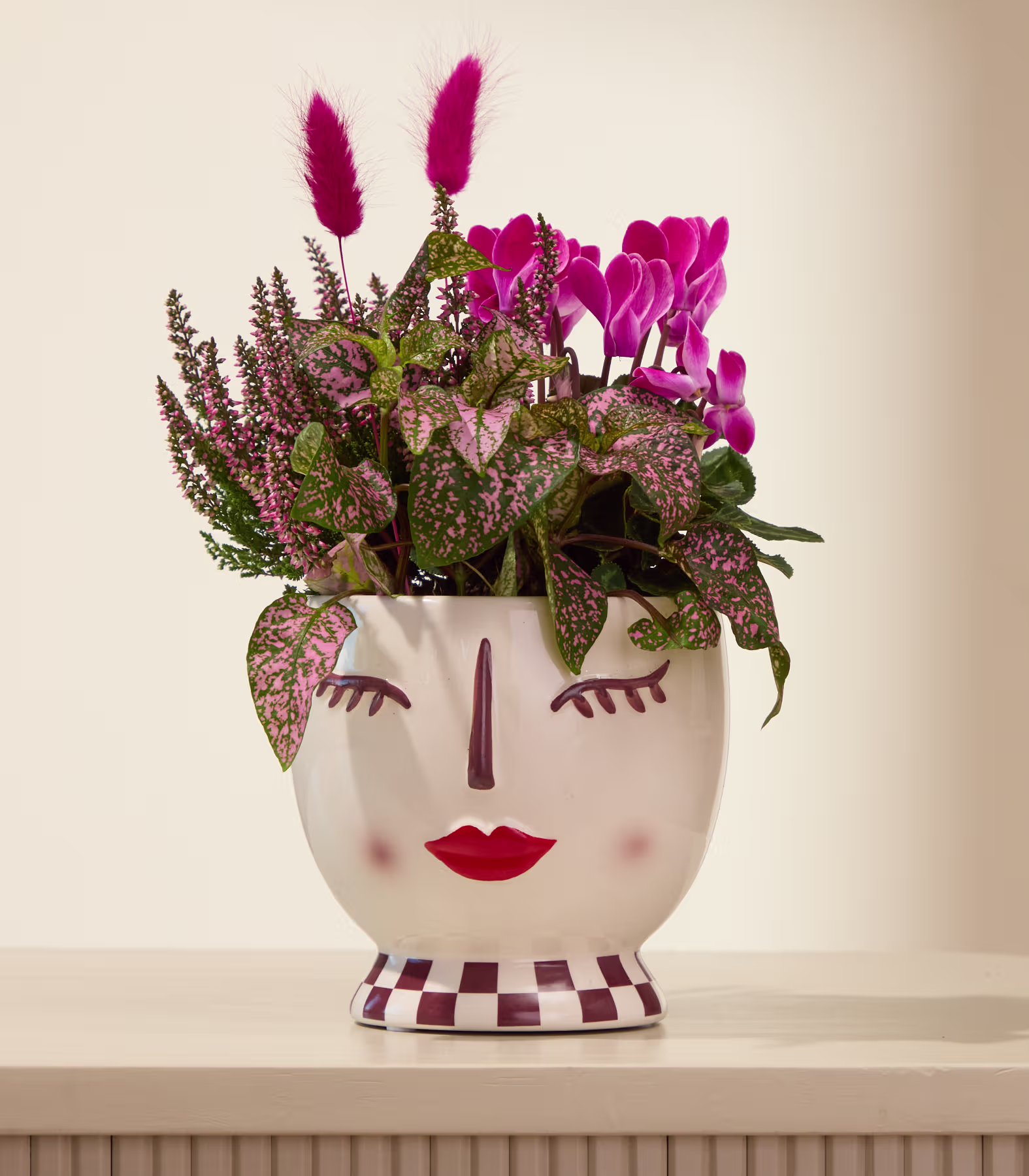 Ein farbenfroher Strauß aus rosa Tulpen, Heidekraut und anderen Blumen, arrangiert in einer gepunkteten Keramikvase, steht auf einer hellen Oberfläche vor einem schlichten Hintergrund. (Beschreibungstext KI-generiert)