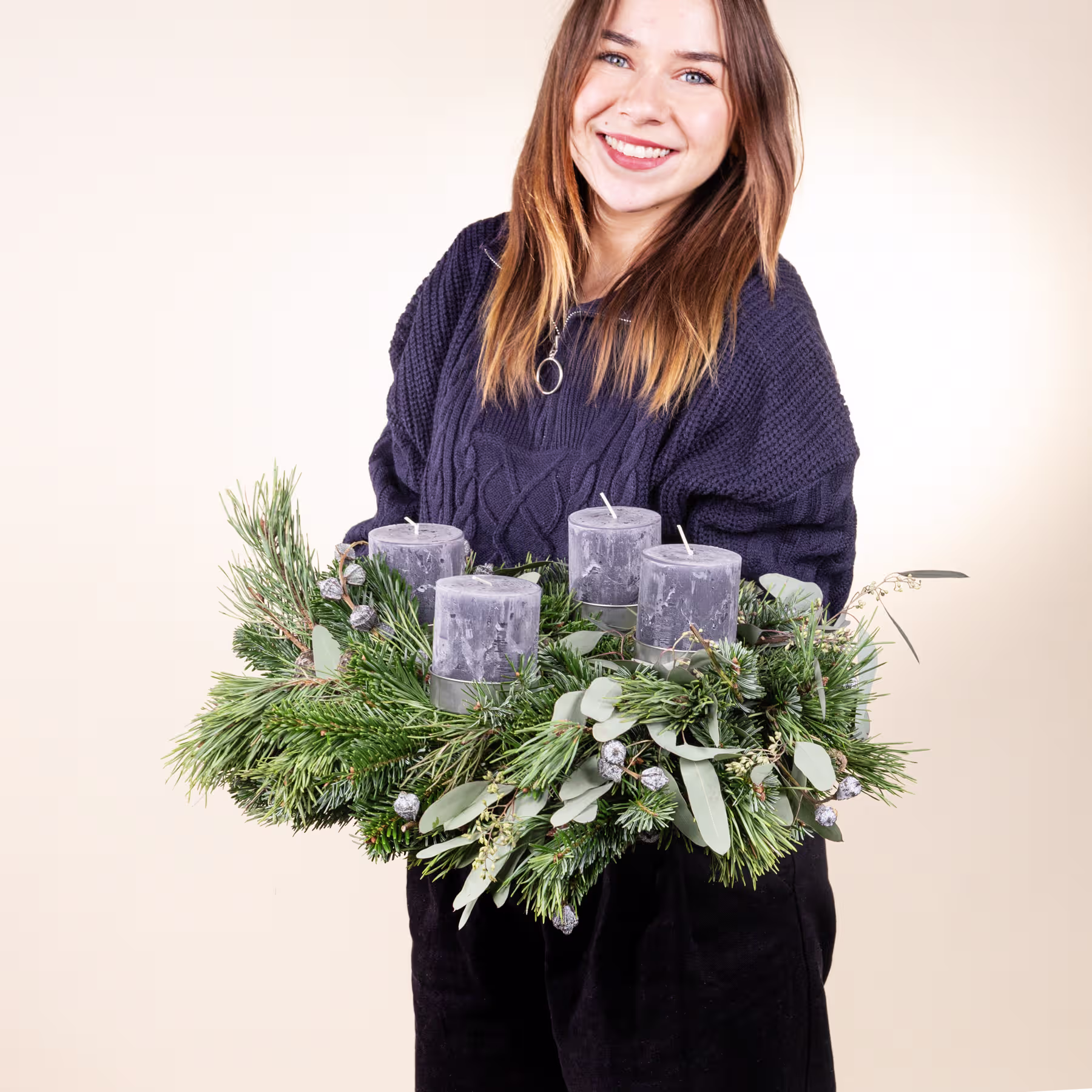 Eine junge Frau mit langen braunen Haaren lächelt und hält vor einem schlichten Hintergrund ein großes Blumenarrangement mit Kerzen, Tannenzweigen und anderem Grün. (Beschreibungstext KI-generiert)