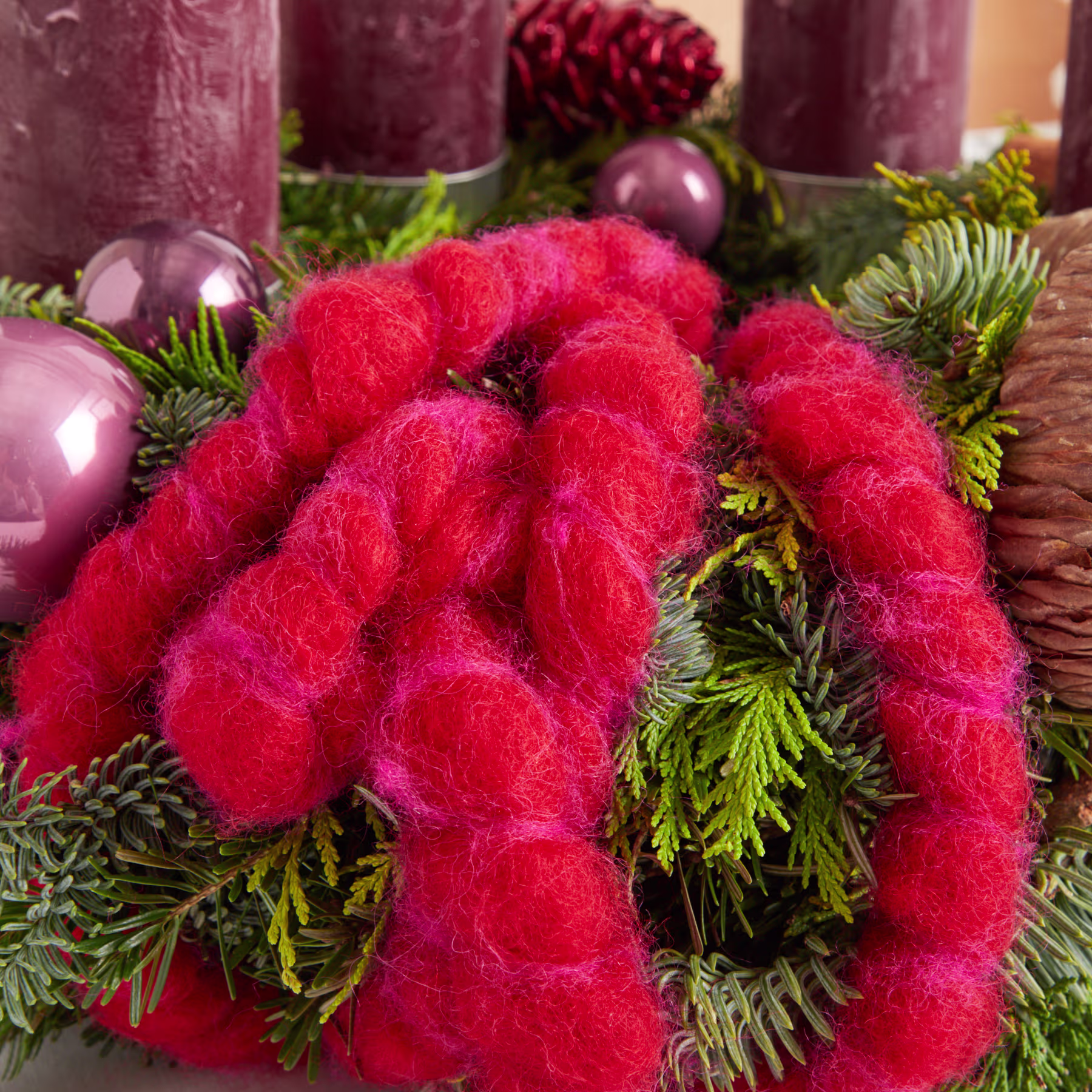 Das Bild zeigt eine leuchtend rote Blume mit flauschiger Textur, umgeben von immergrünem Laub, Tannenzapfen und violetten Ornamenten, wodurch ein festliches und saisonales Arrangement entsteht. (Beschreibungstext KI-generiert)