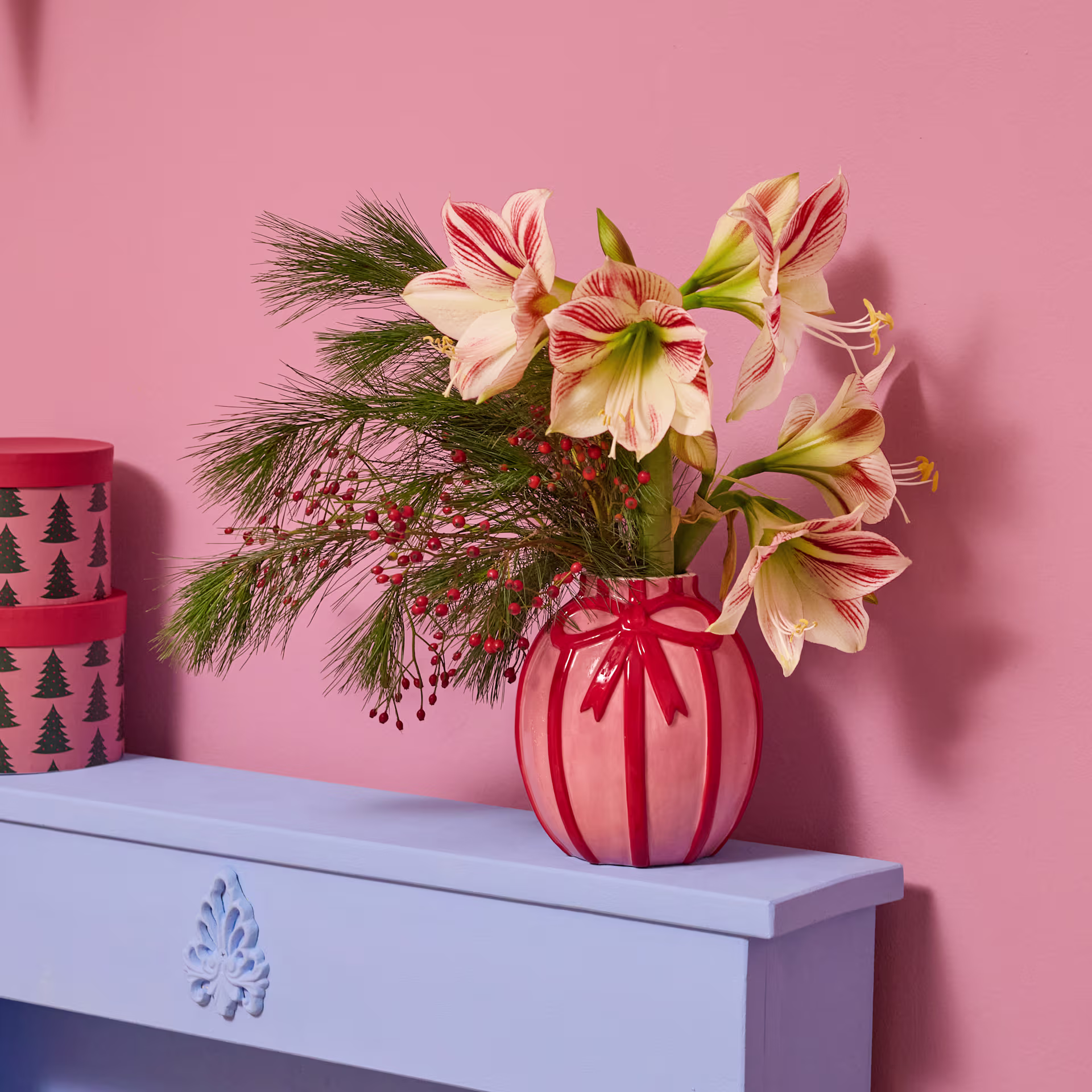 Ein leuchtender Strauß rosa und gelber Blumen steht in einer roten Vase auf einem blauen Tisch vor einem rosa Hintergrund, im Hintergrund ist ein gemusterter Behälter zu sehen. (Beschreibungstext KI-generiert)