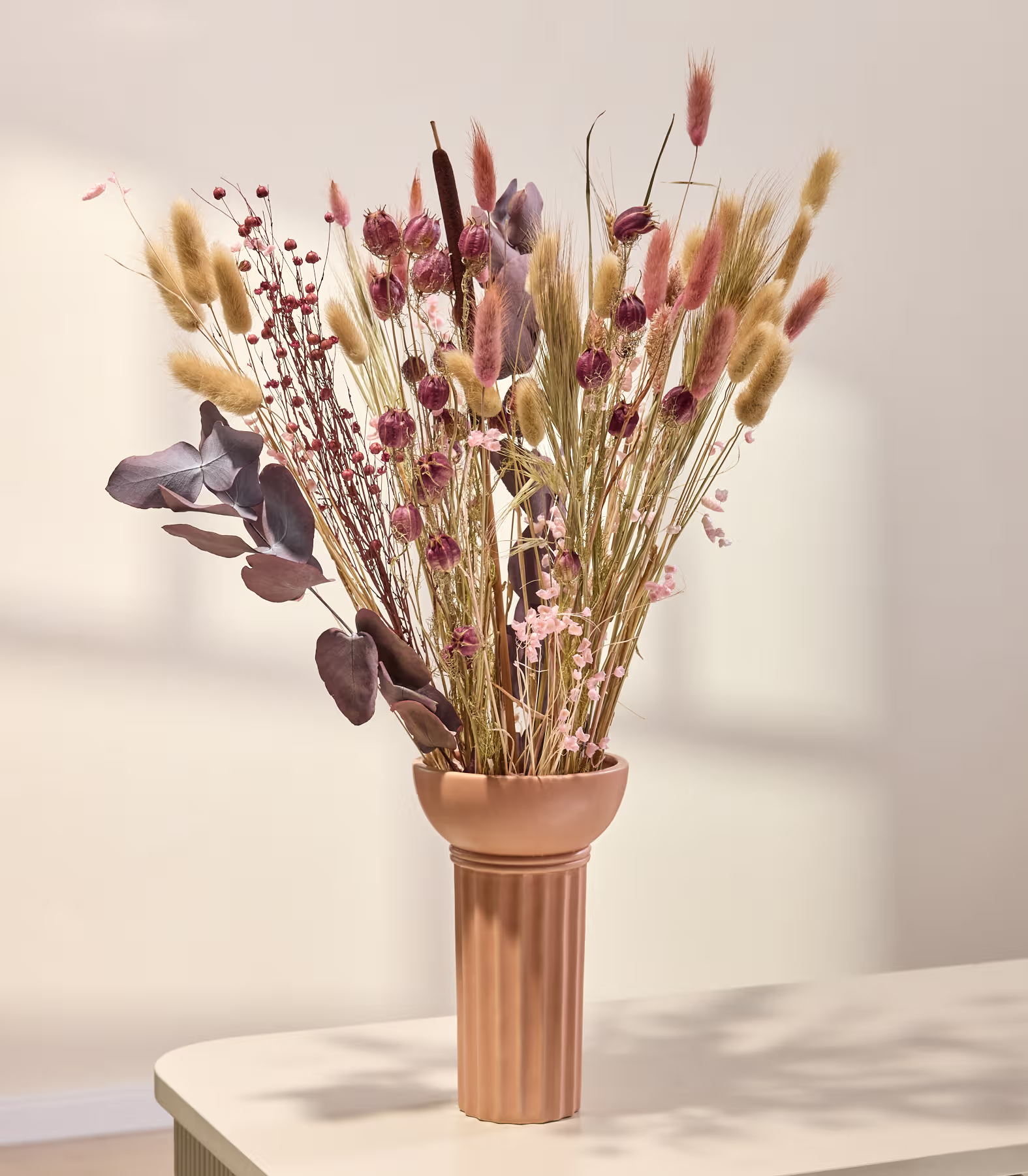 Ein Strauß getrockneter Blumen und Blätter in einer Kupfervase steht auf einem Holztisch vor einem schlichten Hintergrund. (Beschreibungstext KI-generiert)