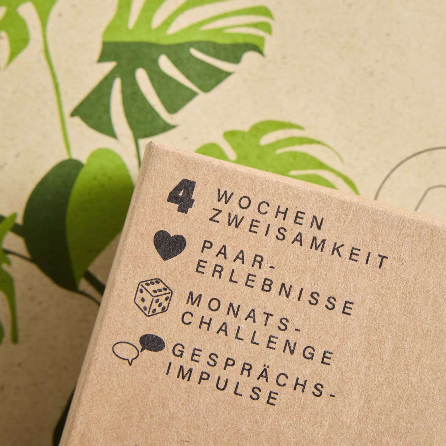 Das Bild zeigt eine braune Papierkarte mit deutschem Text, darunter die Worte „Wochen Zweisam-keit“, „Paar-Erlebnisse“ und „Monats-Challenge“ sowie verschiedene Icons und Symbole vor einem Hintergrund aus grünen tropischen Blättern. (Beschreibungstext KI-generiert)