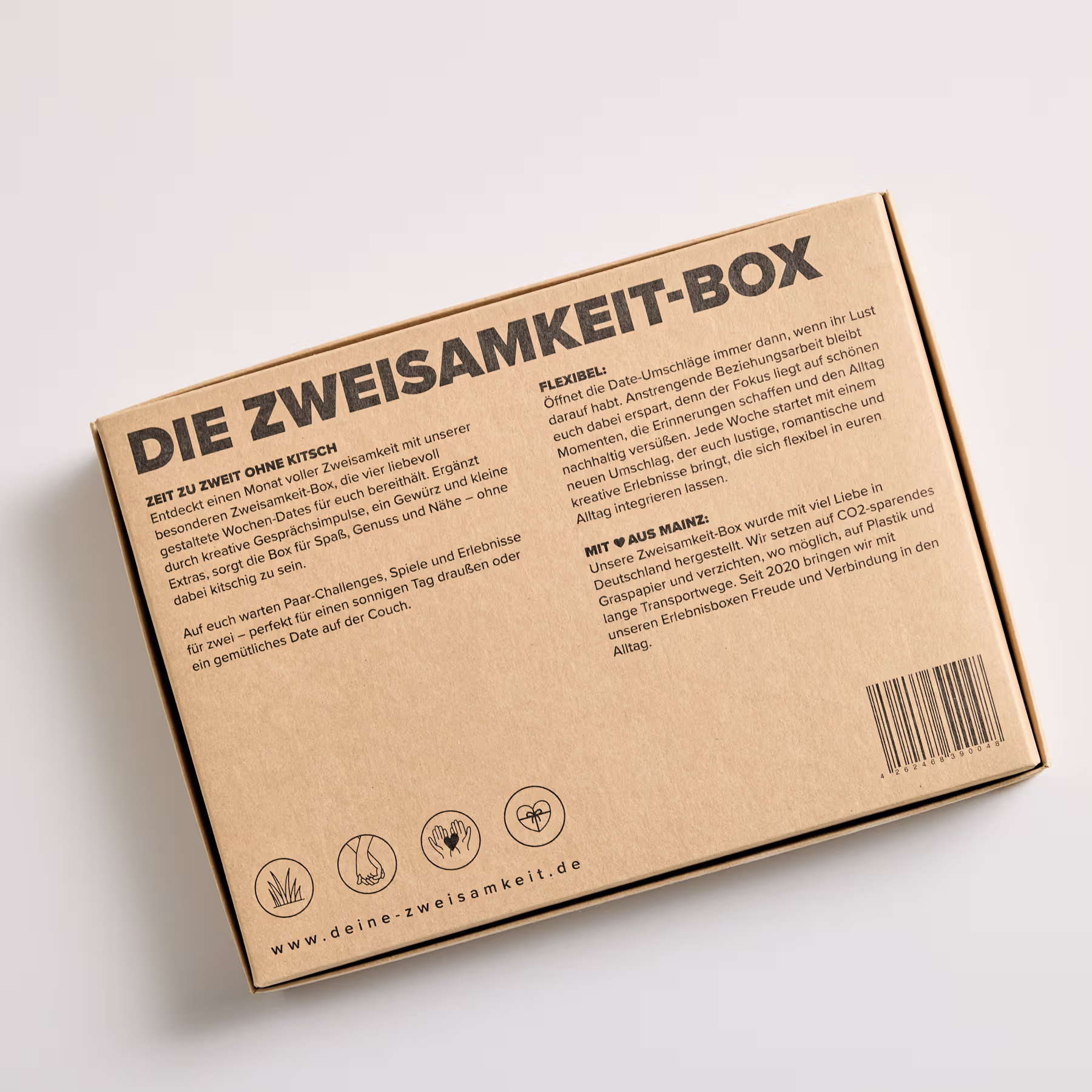 Im Mittelpunkt des Bildes steht ein Karton mit der Aufschrift „Die Zweisamkeit-Box“, der vor einem schlichten weißen Hintergrund steht. (Beschreibungstext KI-generiert)