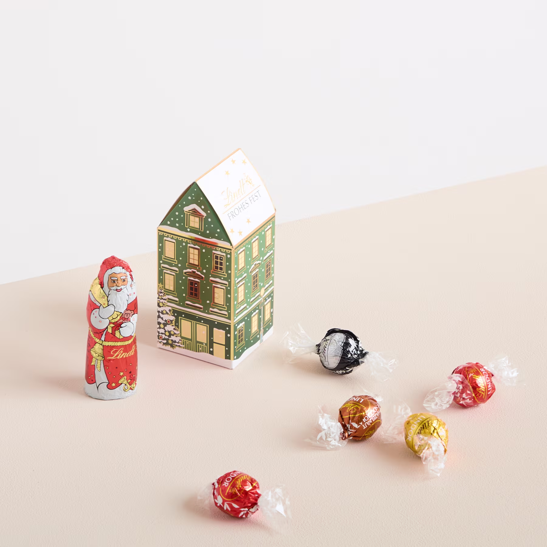 Auf einem beigen Hintergrund sind eine kleine, hölzerne, hausförmige Dekoration, eine Weihnachtsmannfigur und verschiedene verpackte Süßigkeiten arrangiert. (Beschreibungstext KI-generiert)