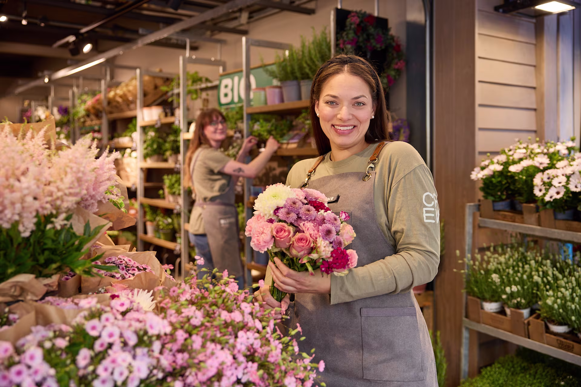 Eine lächelnde Frau in einer geblümten Schürze steht inmitten leuchtender Blumen und Pflanzen in einem Gebäude, das wie ein Blumenladen oder eine Gärtnerei aussieht. (Beschreibungstext KI-generiert)