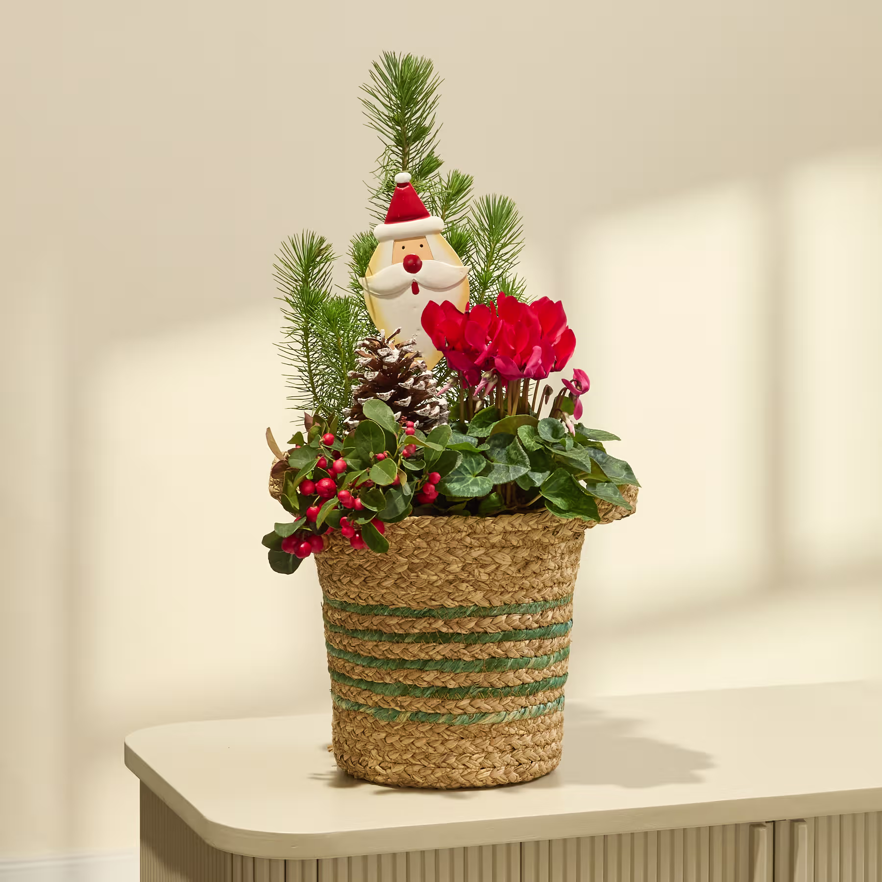 Ein festliches Weihnachtsarrangement in einem geflochtenen Korb, bestehend aus einer kleinen Schneemannfigur, Kiefernzweigen, roten Beeren und anderem saisonalem Laub vor einem schlichten weißen Hintergrund. (Beschreibungstext KI-generiert)
