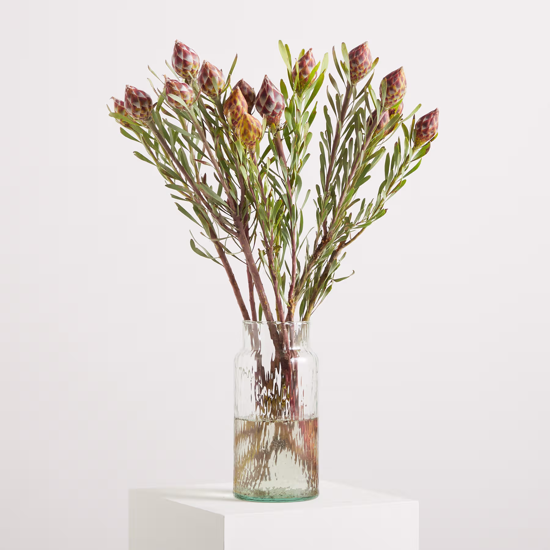 Ein Strauß leuchtender, bunter Blumen in einer Glasvase steht auf einer schlichten weißen Fläche vor einem neutralen Hintergrund. (Beschreibungstext KI-generiert)