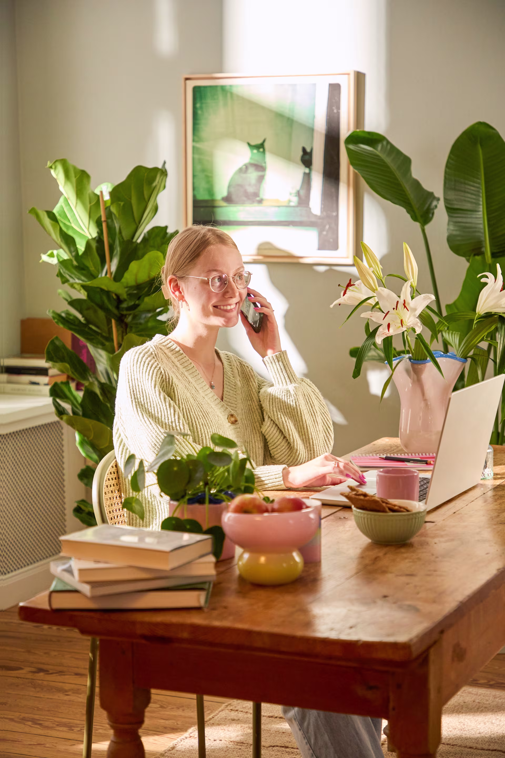 Eine Frau sitzt an einem Holztisch, umgeben von üppigen Zimmerpflanzen, arbeitet an einem Laptop und telefoniert gleichzeitig in einer gemütlichen, pflanzenreichen Wohnatmosphäre. (Beschreibungstext KI-generiert)