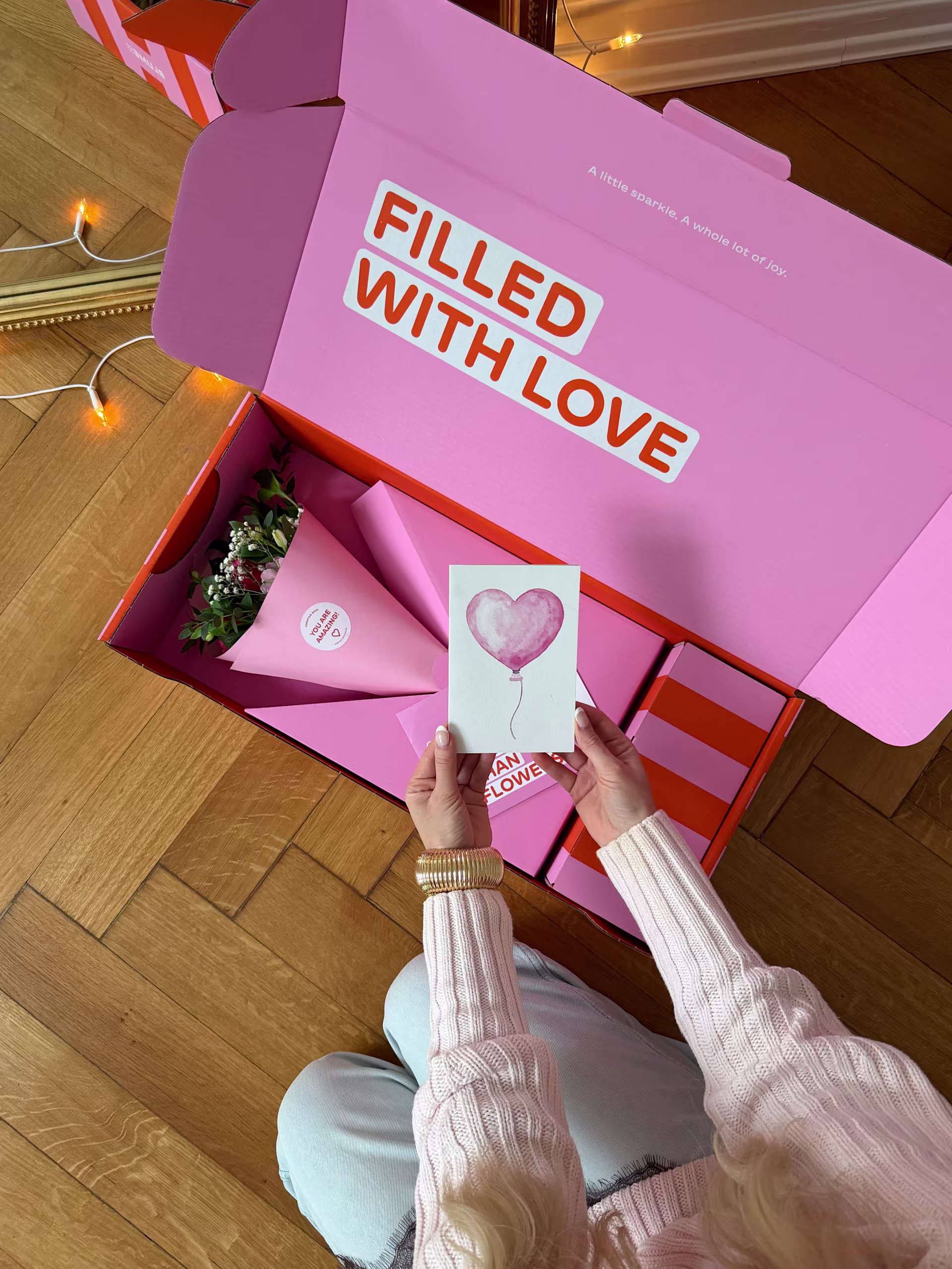 Auf einem Holzboden steht eine große rosa Schachtel mit der Aufschrift „Filled with Love“, in deren Händen eine Karte mit einem Herzsymbol gehalten wird. (Beschreibungstext KI-generiert)