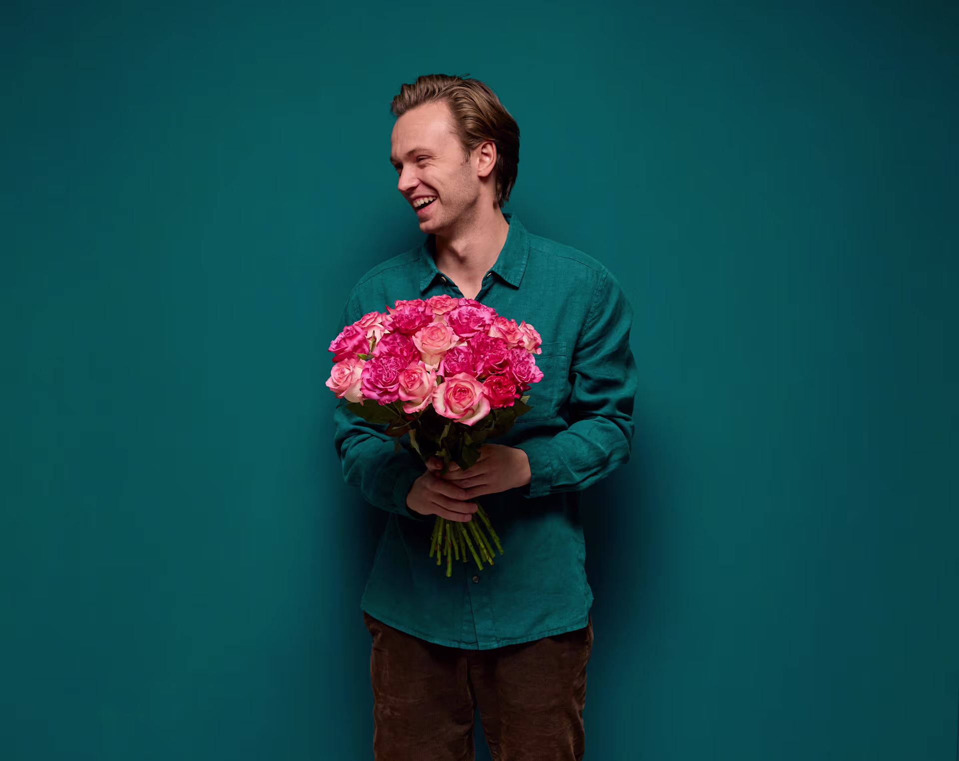 Ein Mann in einem türkisfarbenen Hemd hält einen Strauß leuchtend pinkfarbener Rosen vor einem tief türkisfarbenen Hintergrund. (Beschreibungstext KI-generiert)