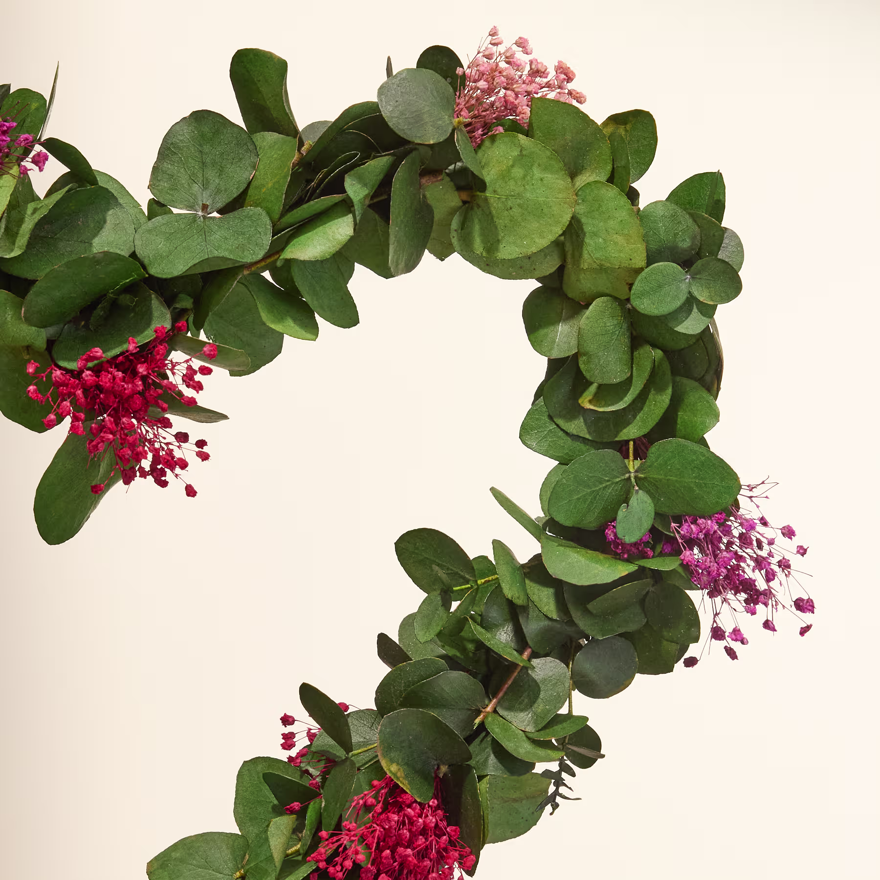 Ein herzförmiger Kranz aus üppigem Grün und leuchtend rosa Blumen wird vor einer Person gehalten, die einen lila Pullover vor einem schlichten Hintergrund trägt. (Beschreibungstext KI-generiert)