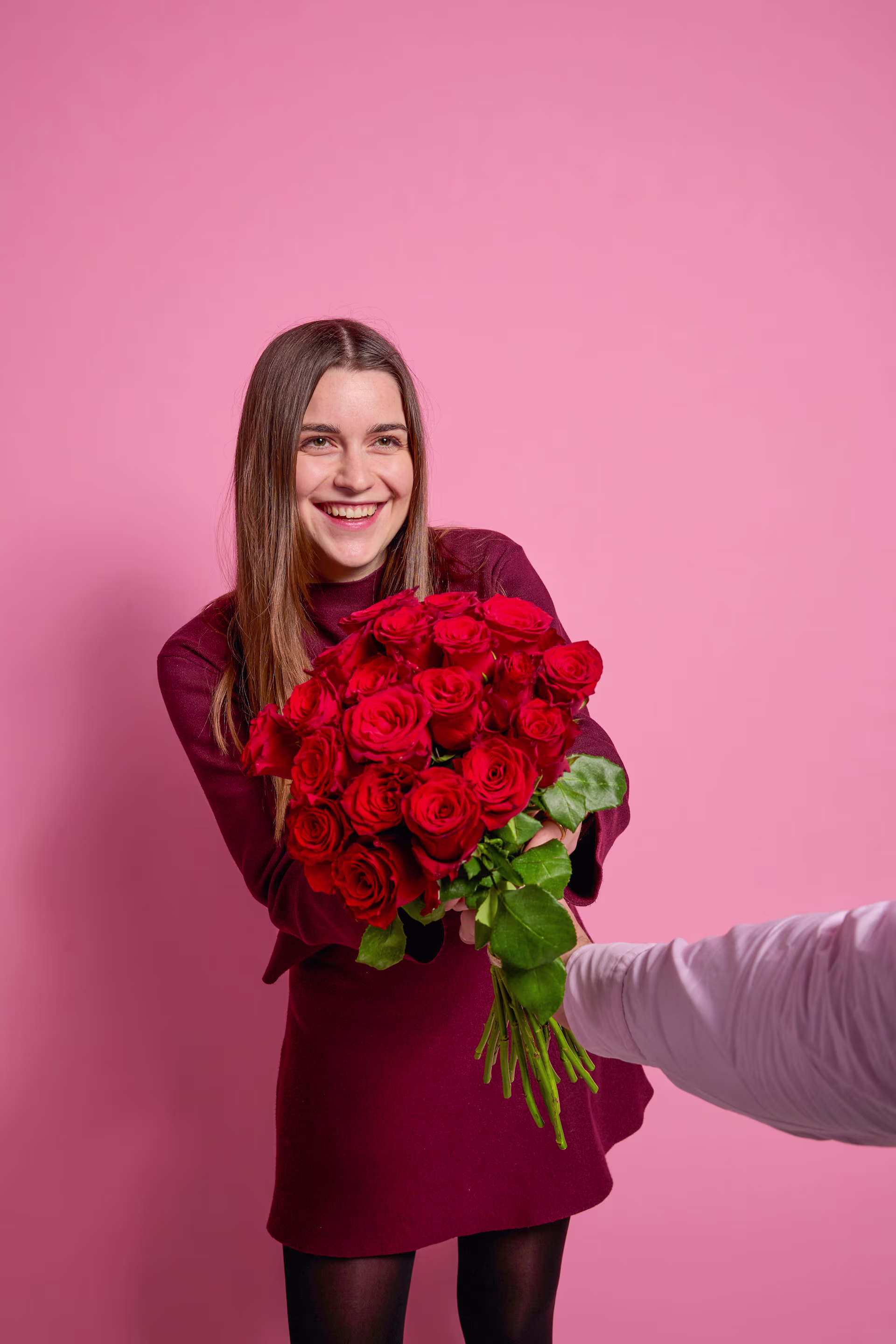 Eine lächelnde junge Frau in einem bordeauxroten Mantel hält einen großen Strauß roter Rosen vor einem leuchtend pinkfarbenen Hintergrund. (Beschreibungstext KI-generiert)