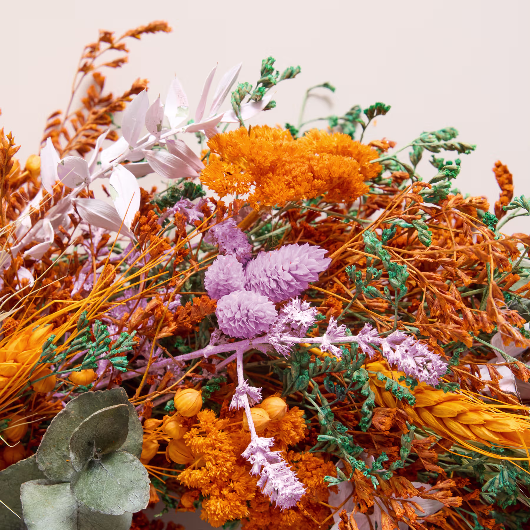 Ein farbenfroher, herbstlicher Kranz aus getrockneten Blumen, Blättern und Gräsern wird vor einem schlichten weißen Hintergrund in einer Hand gehalten. (Beschreibungstext KI-generiert)