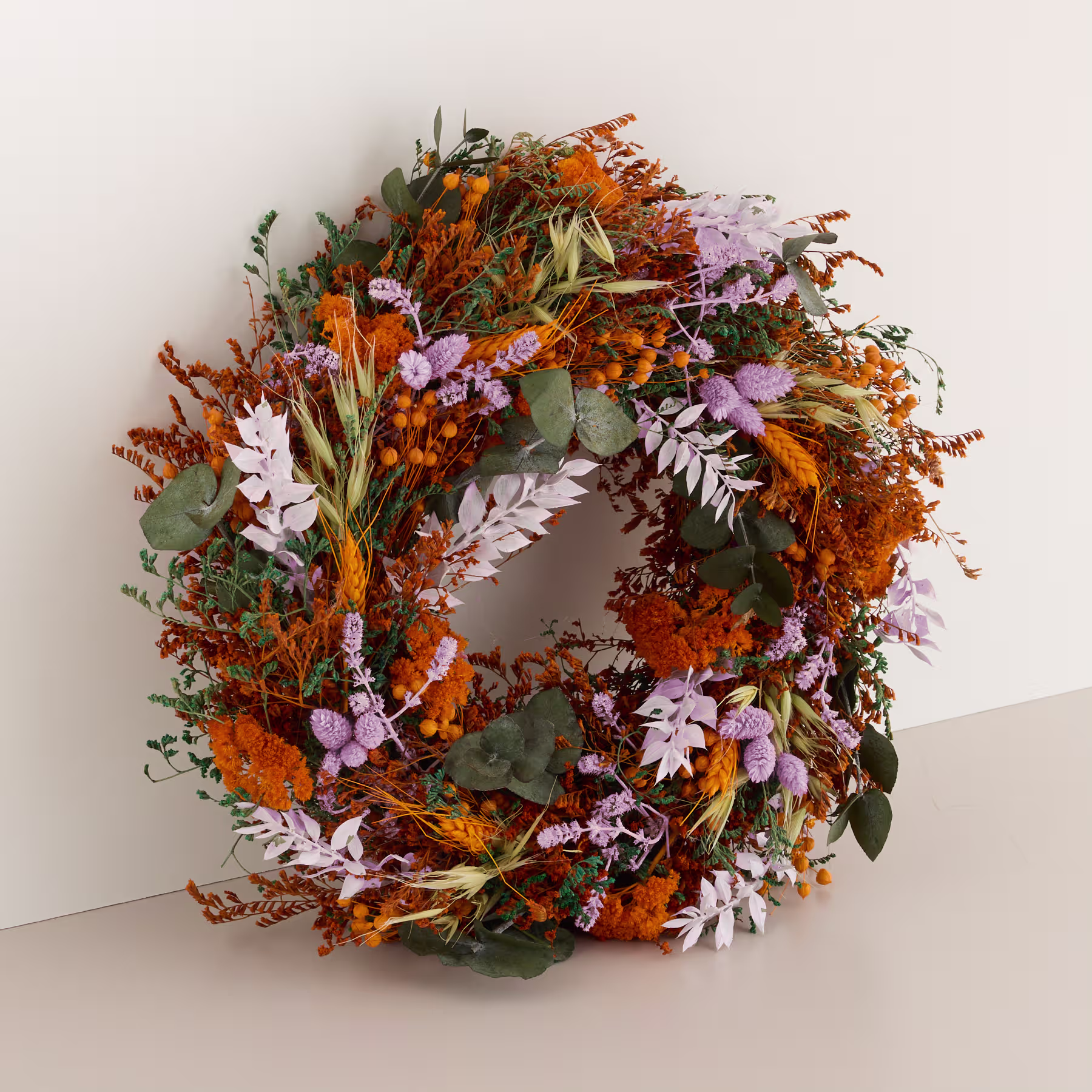 Ein farbenfroher Herbstkranz aus getrockneten Blumen, Blättern und anderen Naturelementen wird von einer Person gehalten, die ein gestreiftes Hemd vor einem schlichten, hellen Hintergrund trägt. (Beschreibungstext KI-generiert)