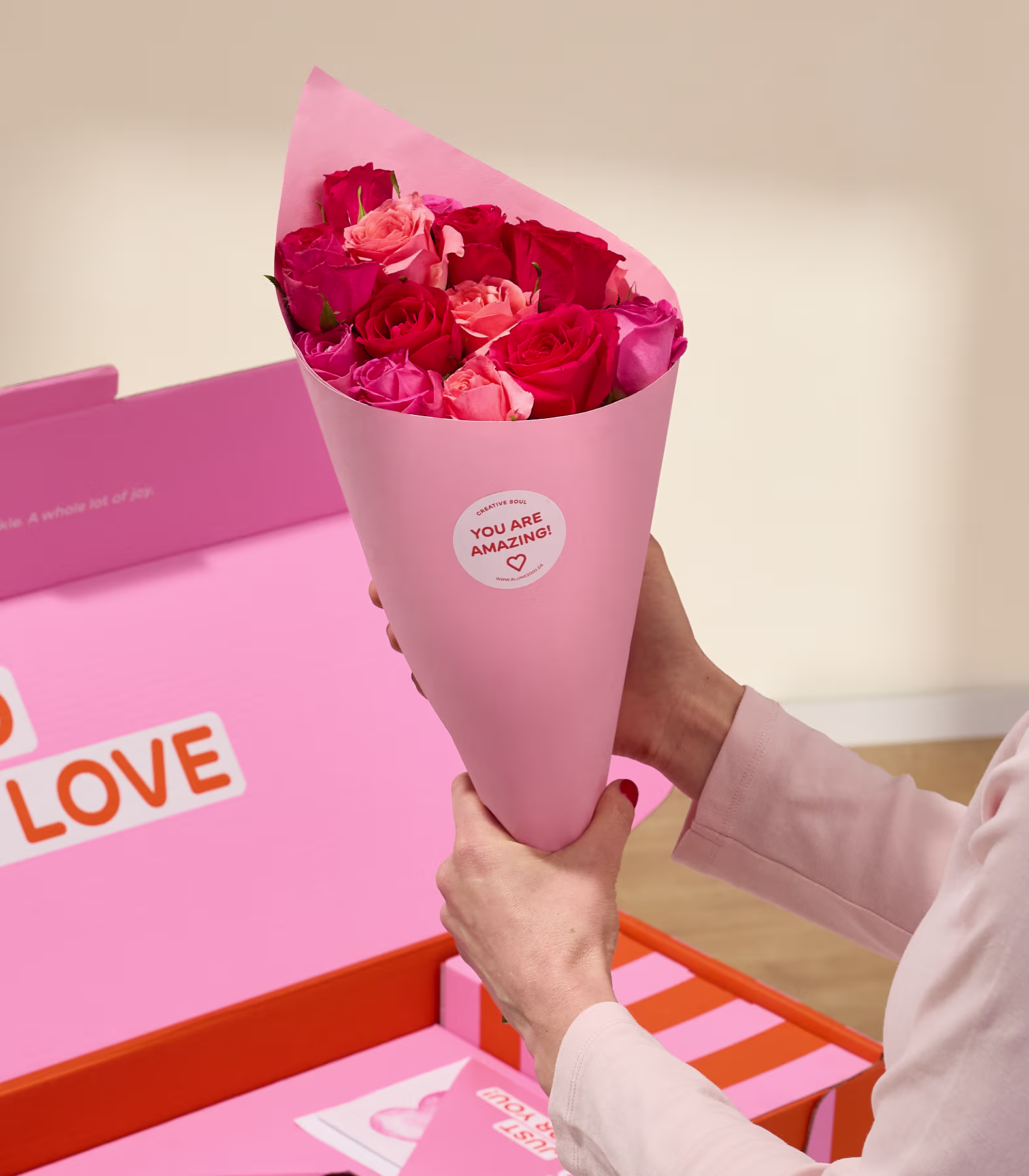 Auf einem Tisch steht eine rosafarbene Geschenkbox mit der Aufschrift „Voller Liebe“, daneben ein Strauß roter Rosen in einer Glasvase. (Beschreibungstext KI-generiert)