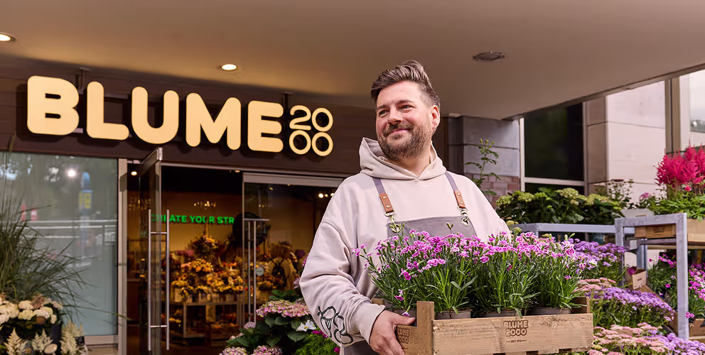 Ein Mann mit Bart und in Freizeitkleidung steht vor einem Blumenladen namens „Blume 2000“ und hält eine Kiste mit bunten Blumen. (Beschreibungstext KI-generiert)