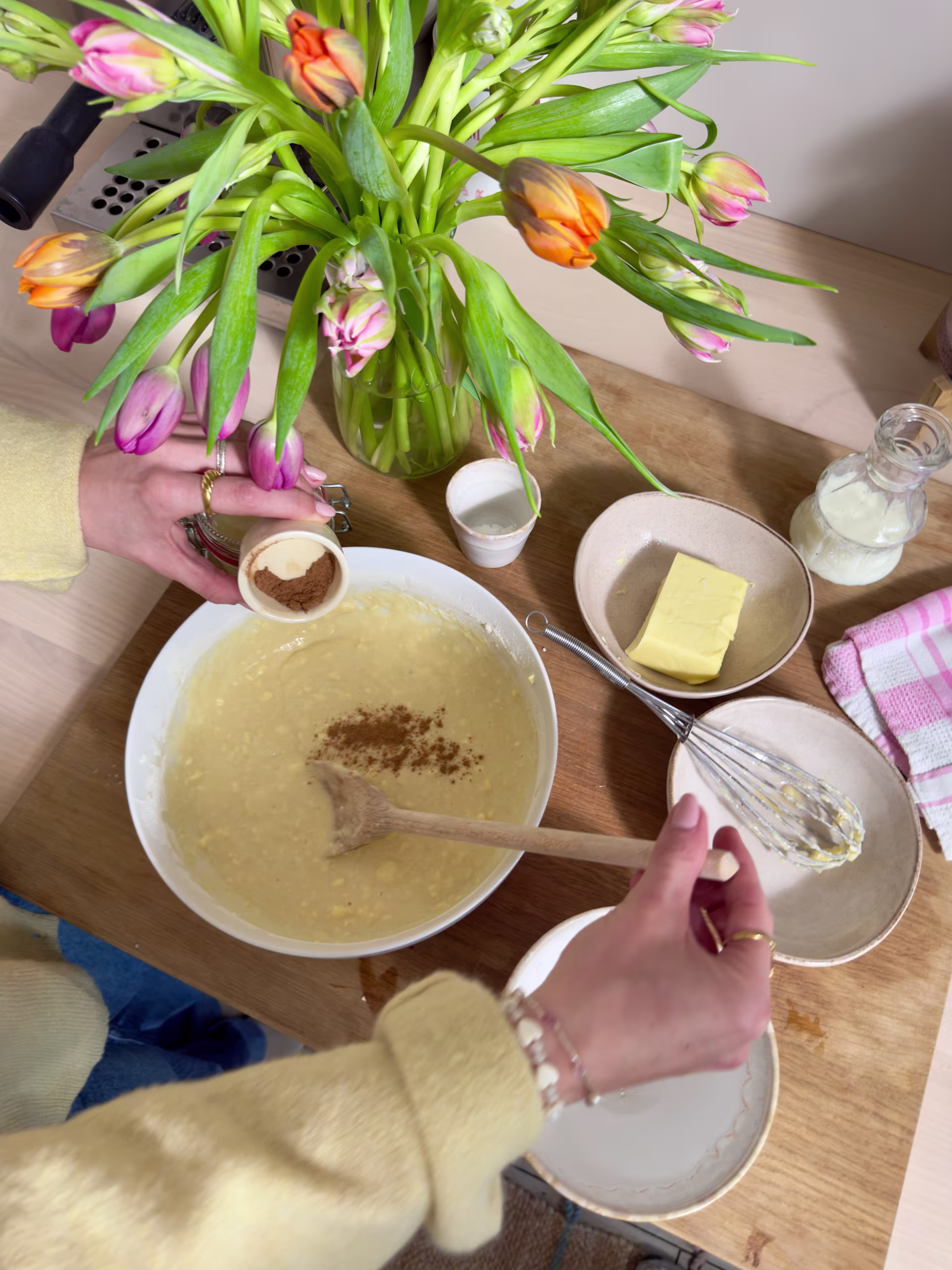 Ein Holztisch mit einer Schüssel Suppe, Butter und einer Vase mit bunten Tulpen; im Vordergrund sind die Hände einer Person zu sehen. (Beschreibungstext KI-generiert)