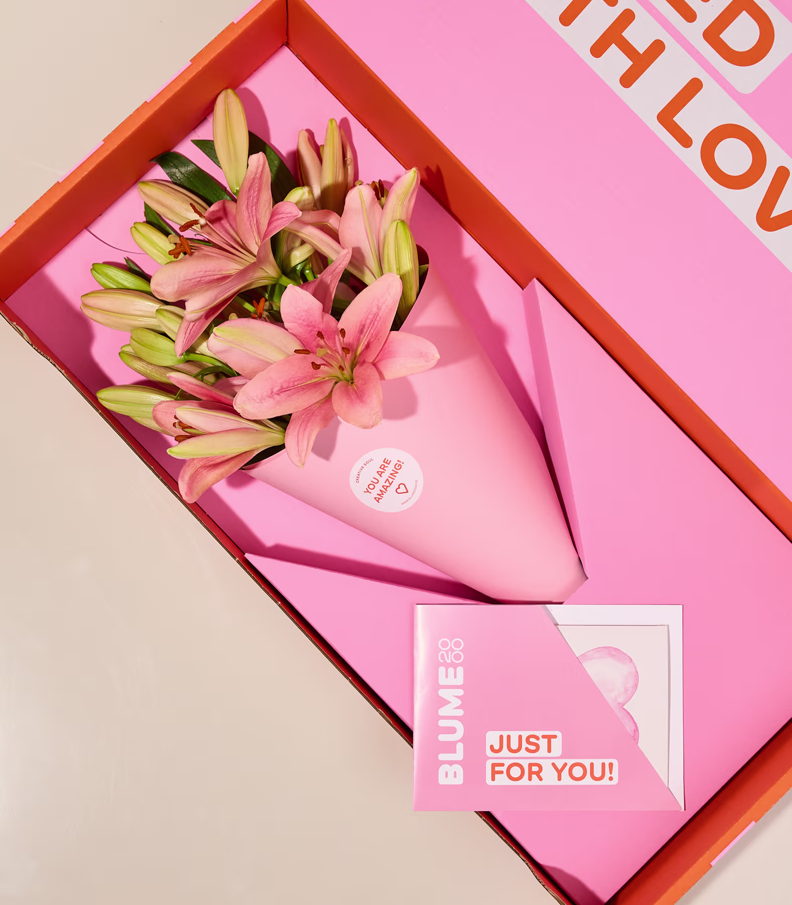 In einer roten Schachtel wird ein Strauß rosa und gelber Lilien präsentiert, dazu eine Grußkarte mit dem Text „Nur für dich!“ vor hellem Hintergrund. (Beschreibungstext KI-generiert)